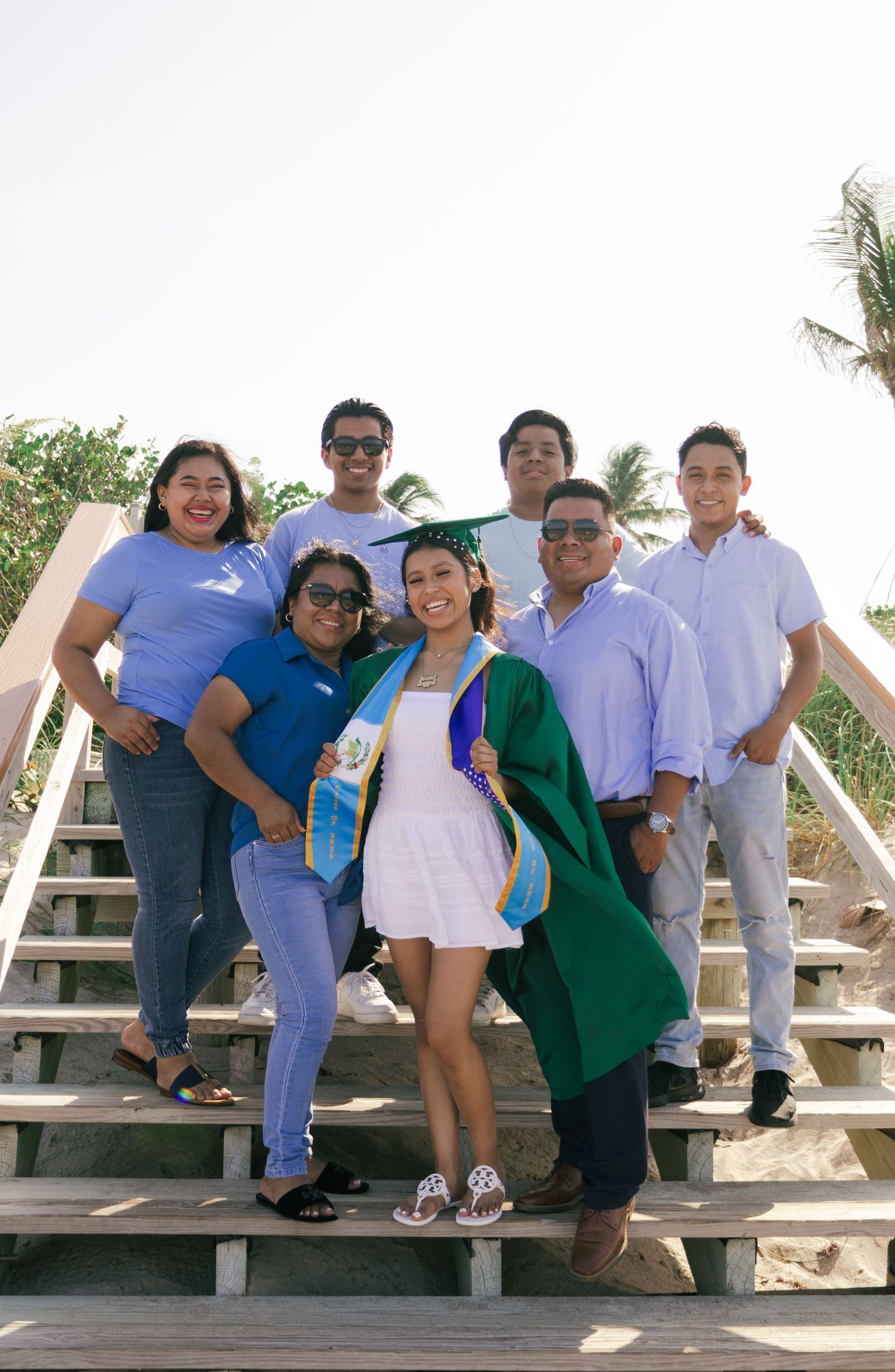 Family celebrating graduation on beach stairs, graduate in green cap and gown with blue sash, surrounded by six smiling family members.