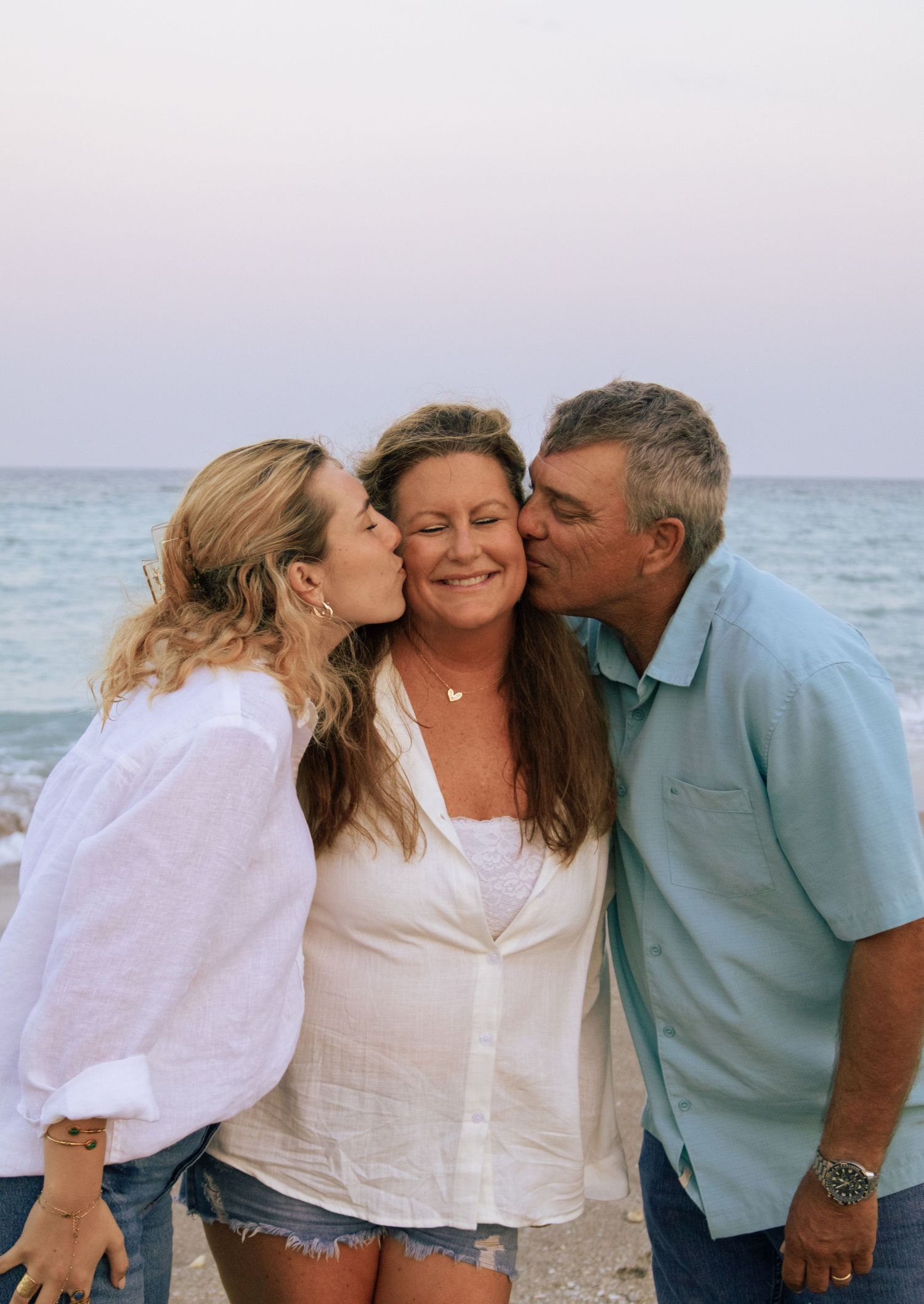 Two people kissing woman on cheeks simultaneously on beach at sunset in joyful family moment.