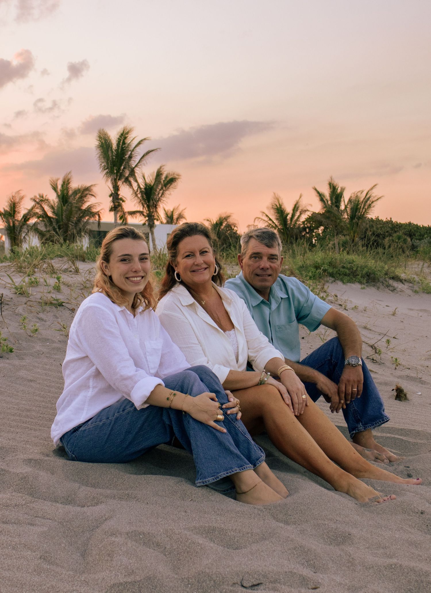 Three people sitting together on sandy beach at sunset with palm trees, smiling for a family portrait photo.
