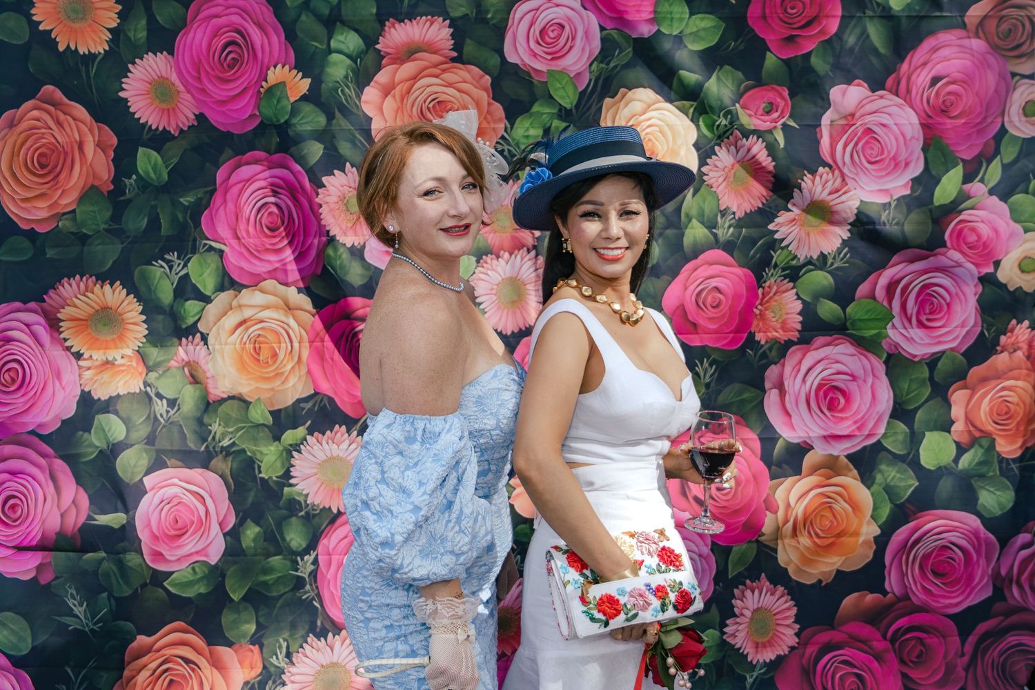 Two women in elegant outfits posing before a vibrant floral rose photo backdrop outdoors.