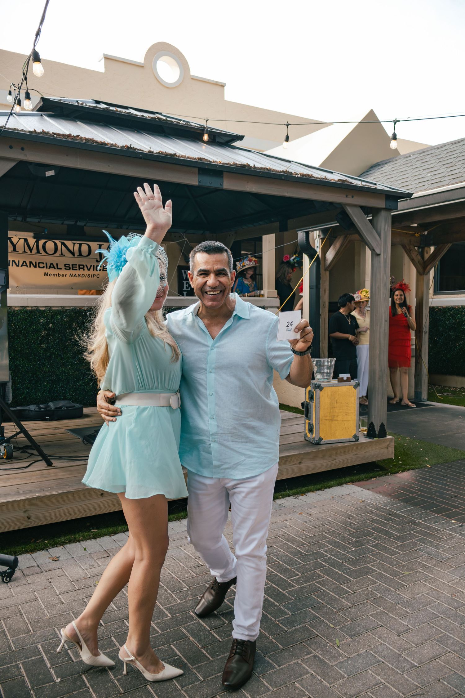 Smiling couple in light blue outfits dancing and waving at outdoor Derby party.