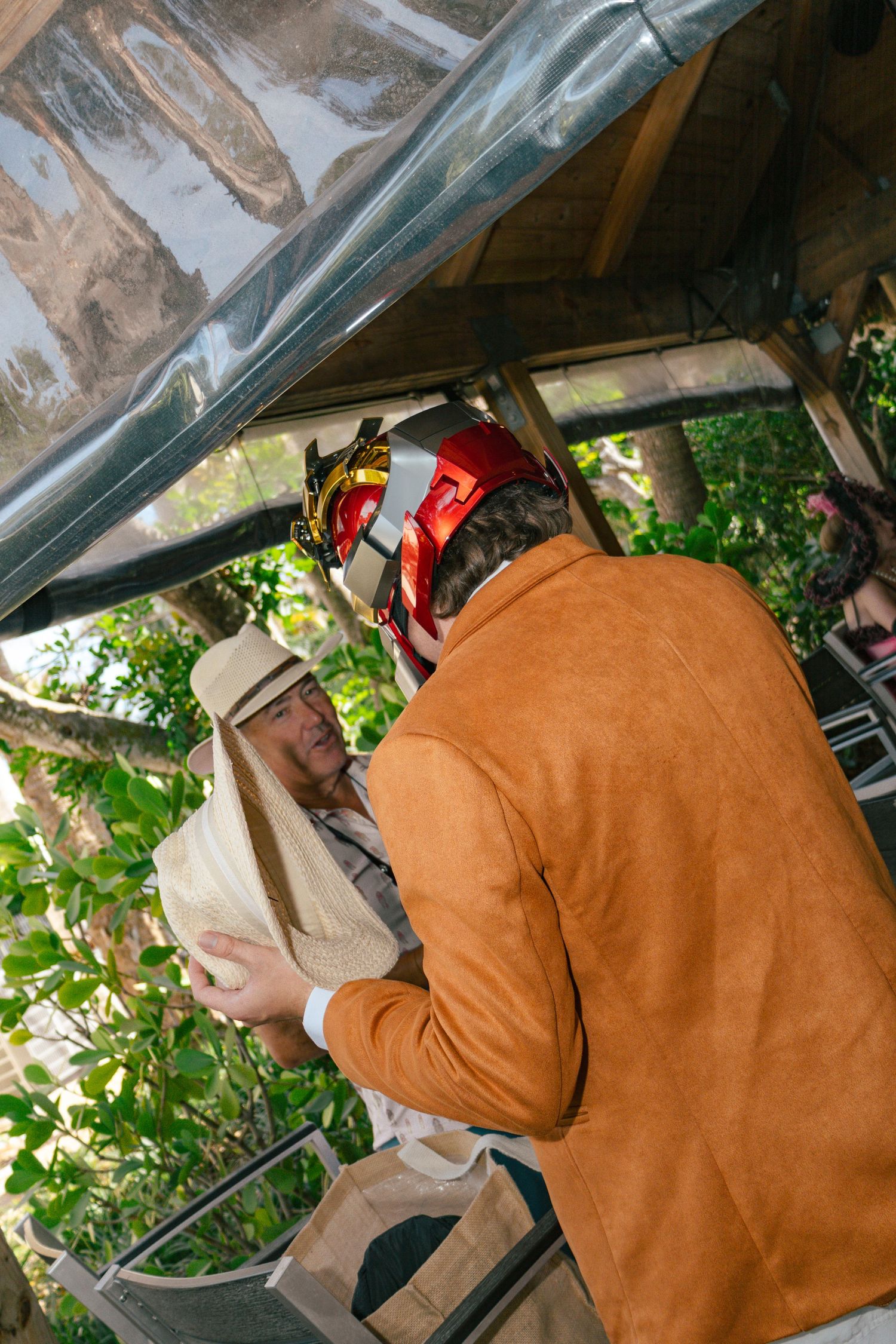 Two people in costumes, one wearing a red helmet, interact under a canopy surrounded by green foliage.