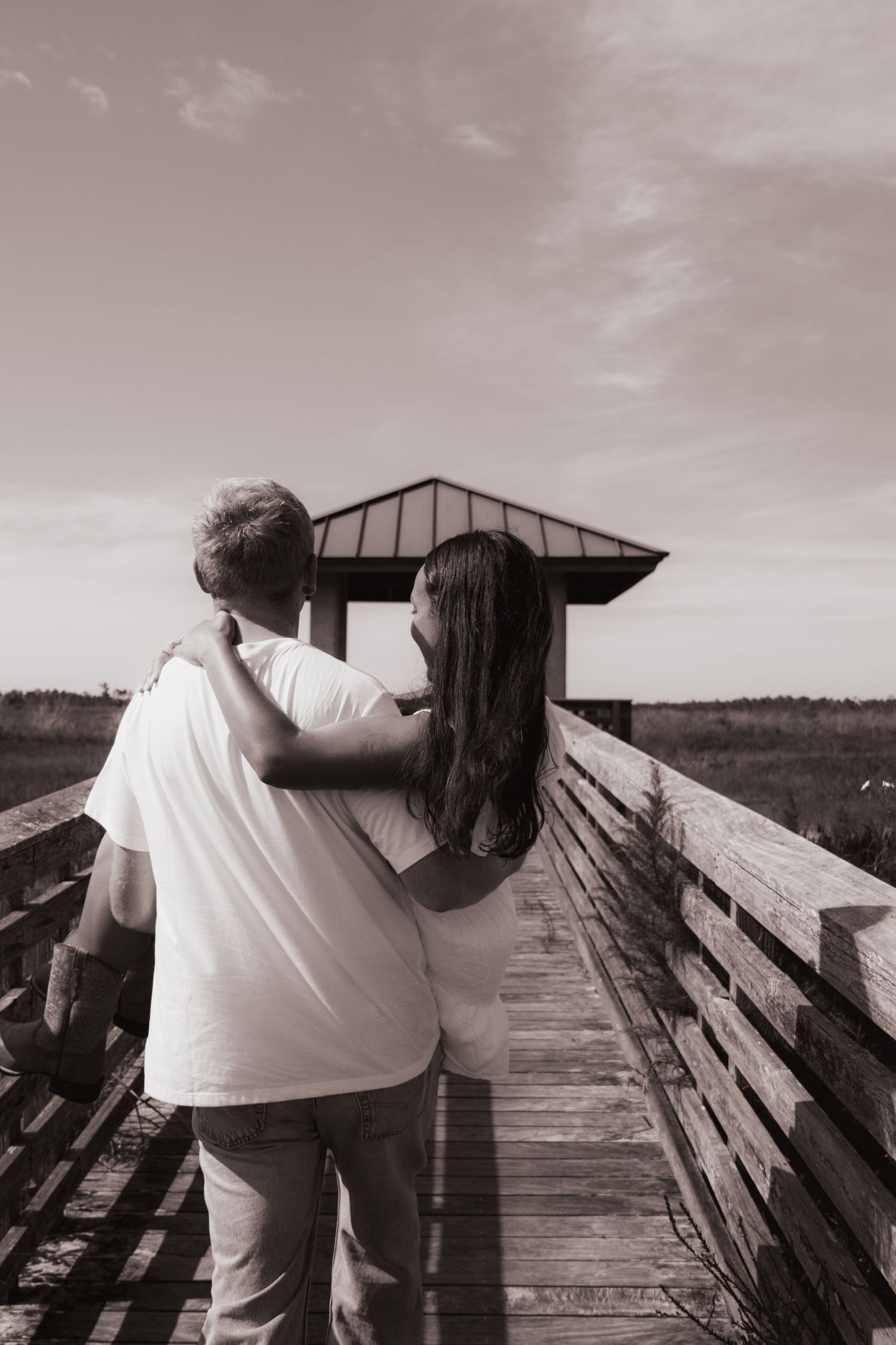 Black and white photo of couple embracing on wooden boardwalk walkway leading to gazebo.
