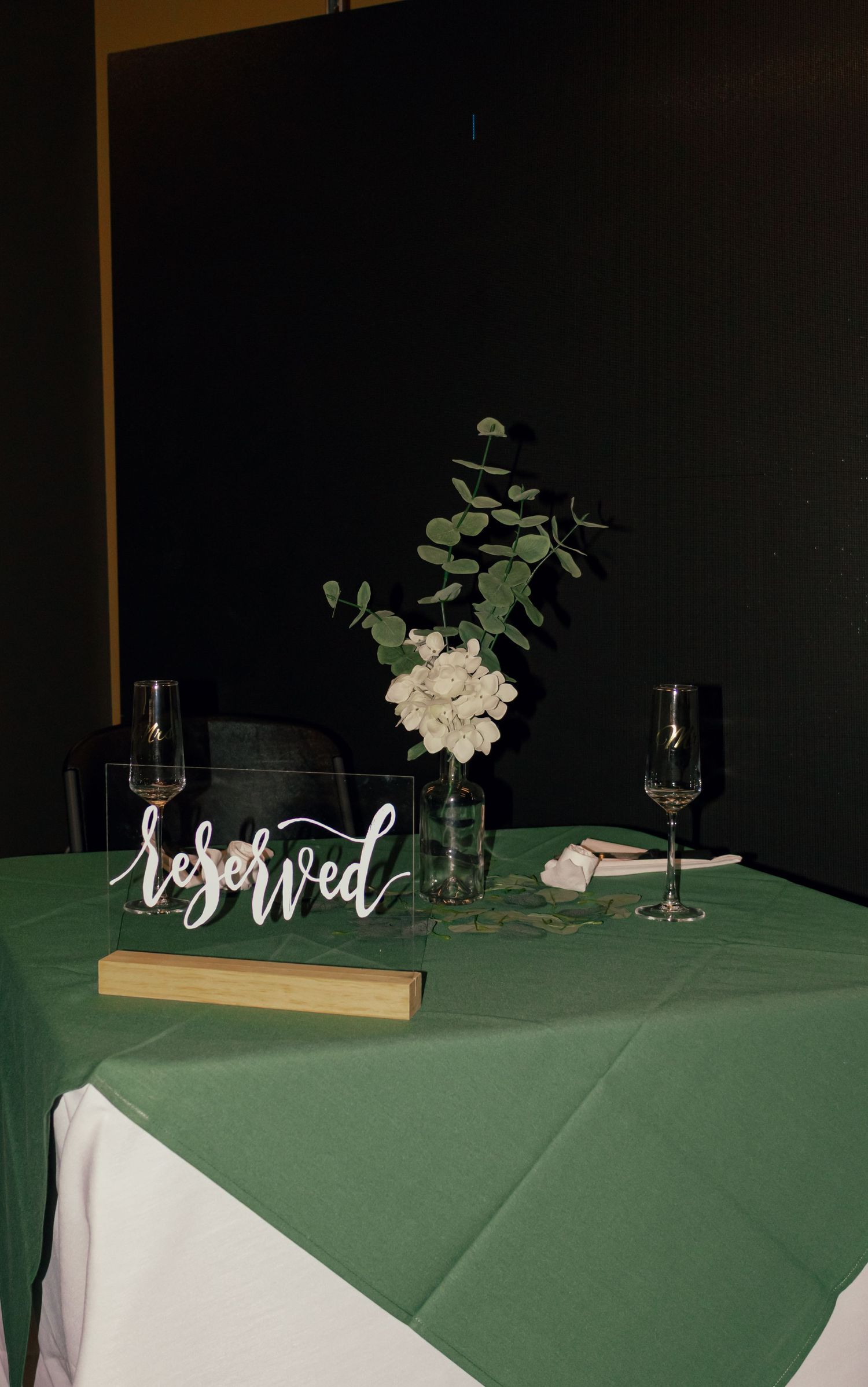 Elegant wedding sweetheart table with green linen, white floral centerpiece, eucalyptus greenery, and acrylic reserved sign.