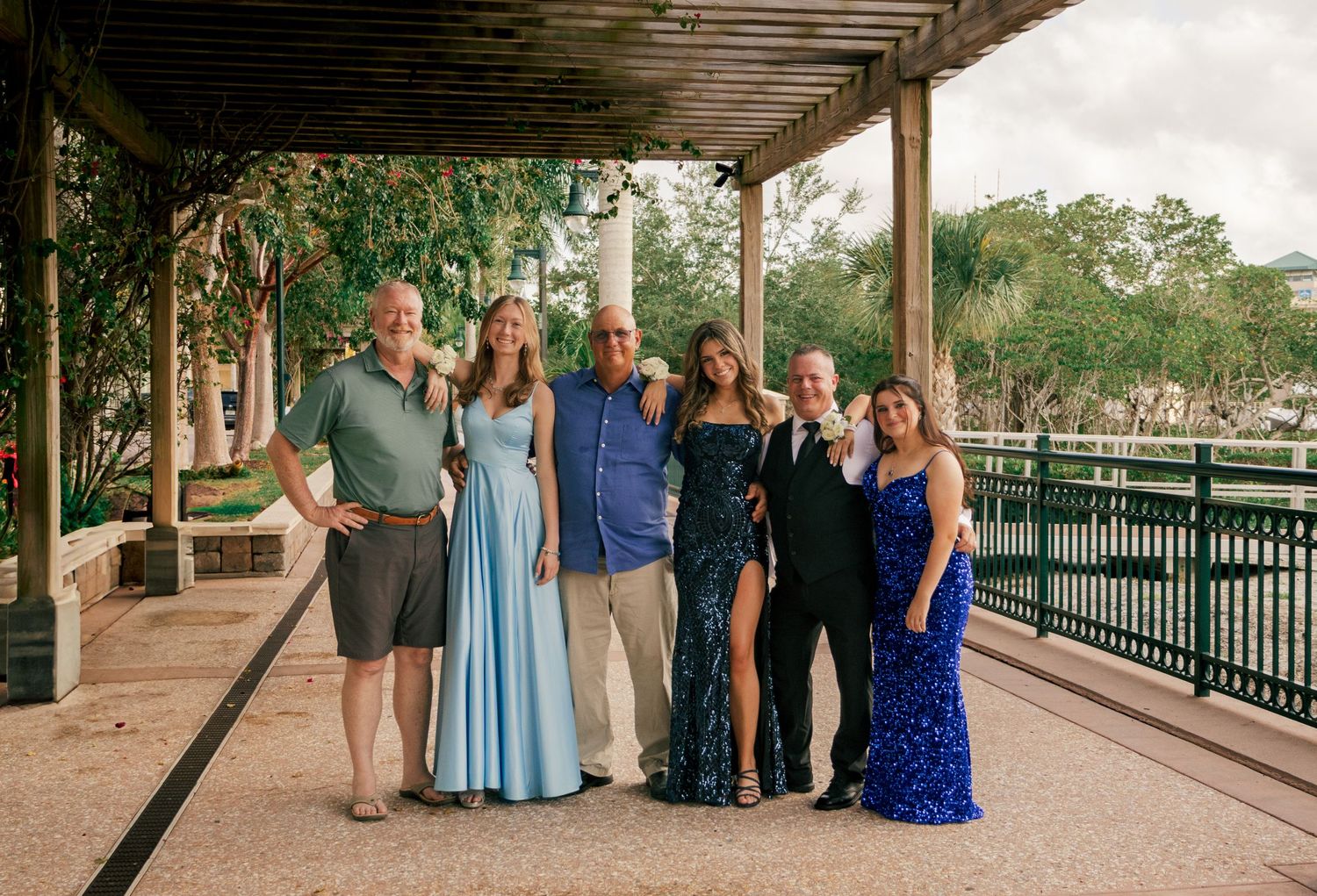 Family group of six adults and teens in formal attire pose under an outdoor pergola walkway.