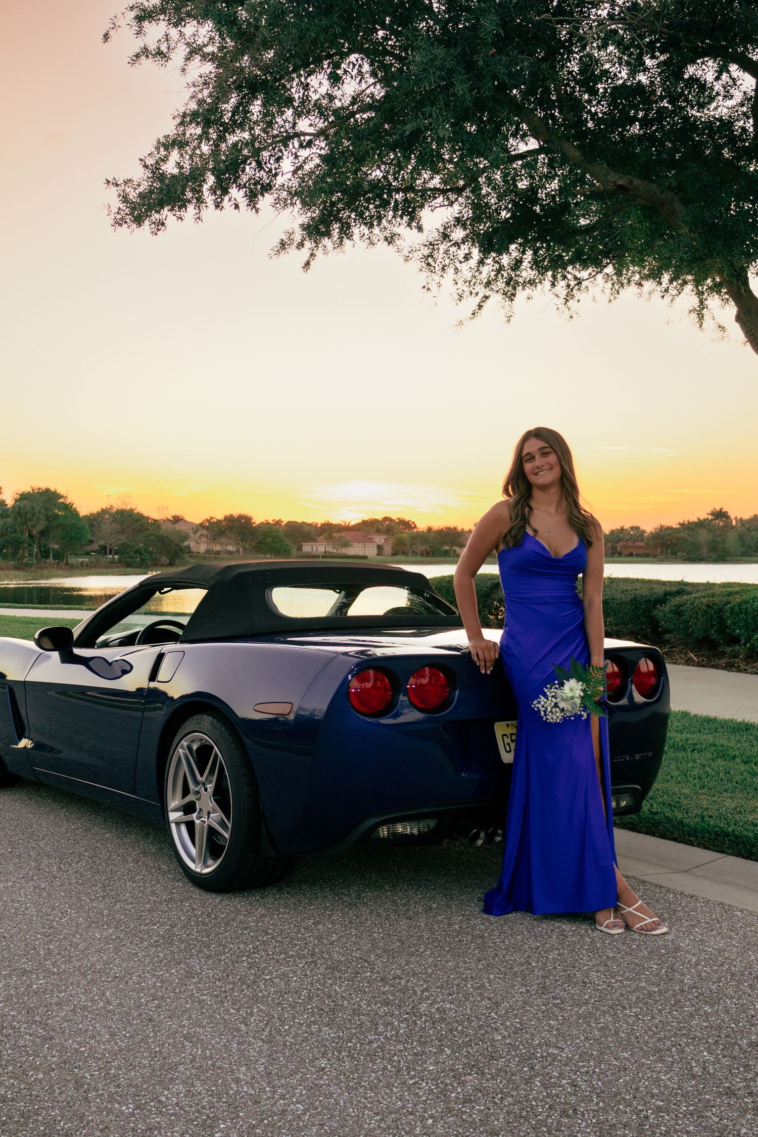 Young woman in a royal blue prom gown posing beside a blue Corvette convertible at sunset.