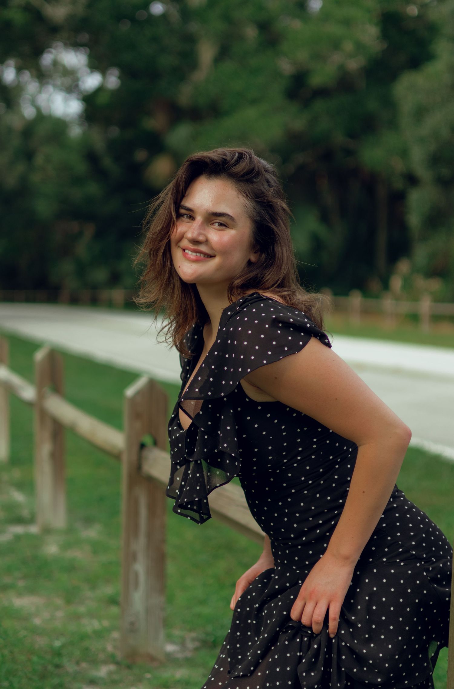 Smiling young woman in black polka dot dress posing near wooden fence in lush green park.