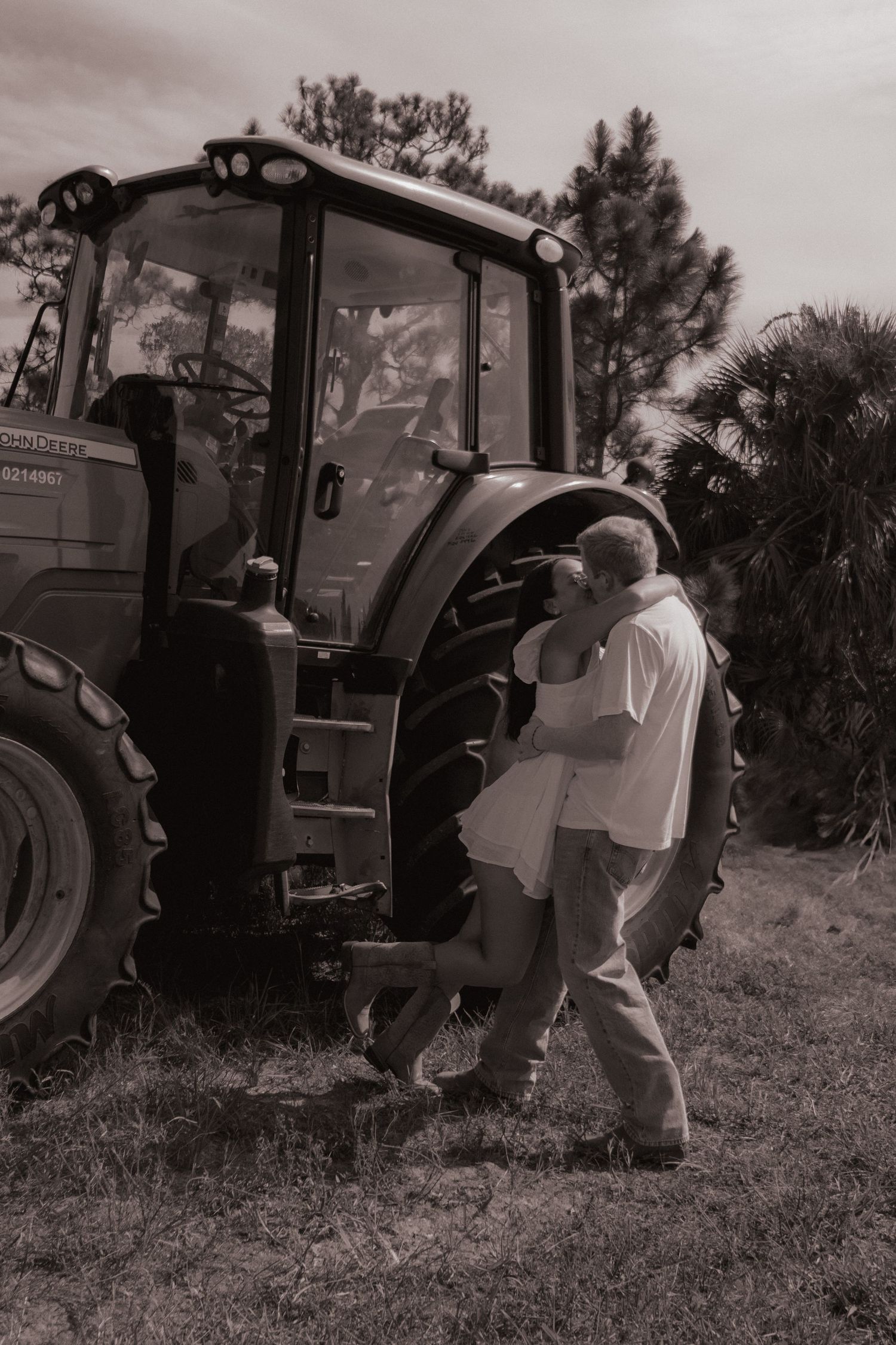 Couple sharing a kiss beside a large green tractor in a sepia-toned outdoor farm setting.