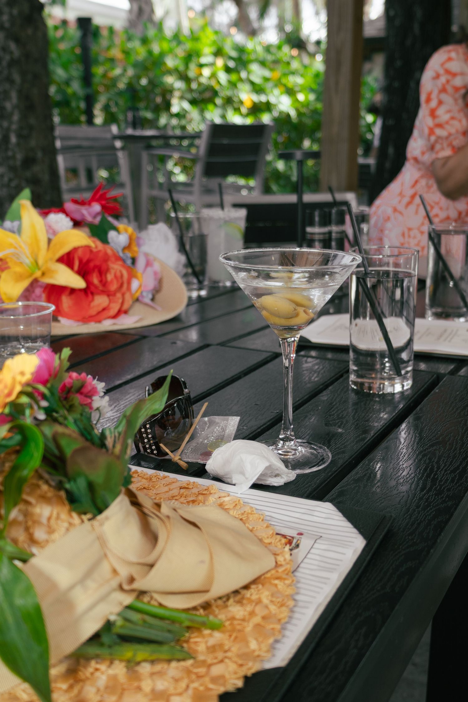 Elegant martini with olives beside fresh tropical flowers and gold napkin on a dark outdoor patio table.