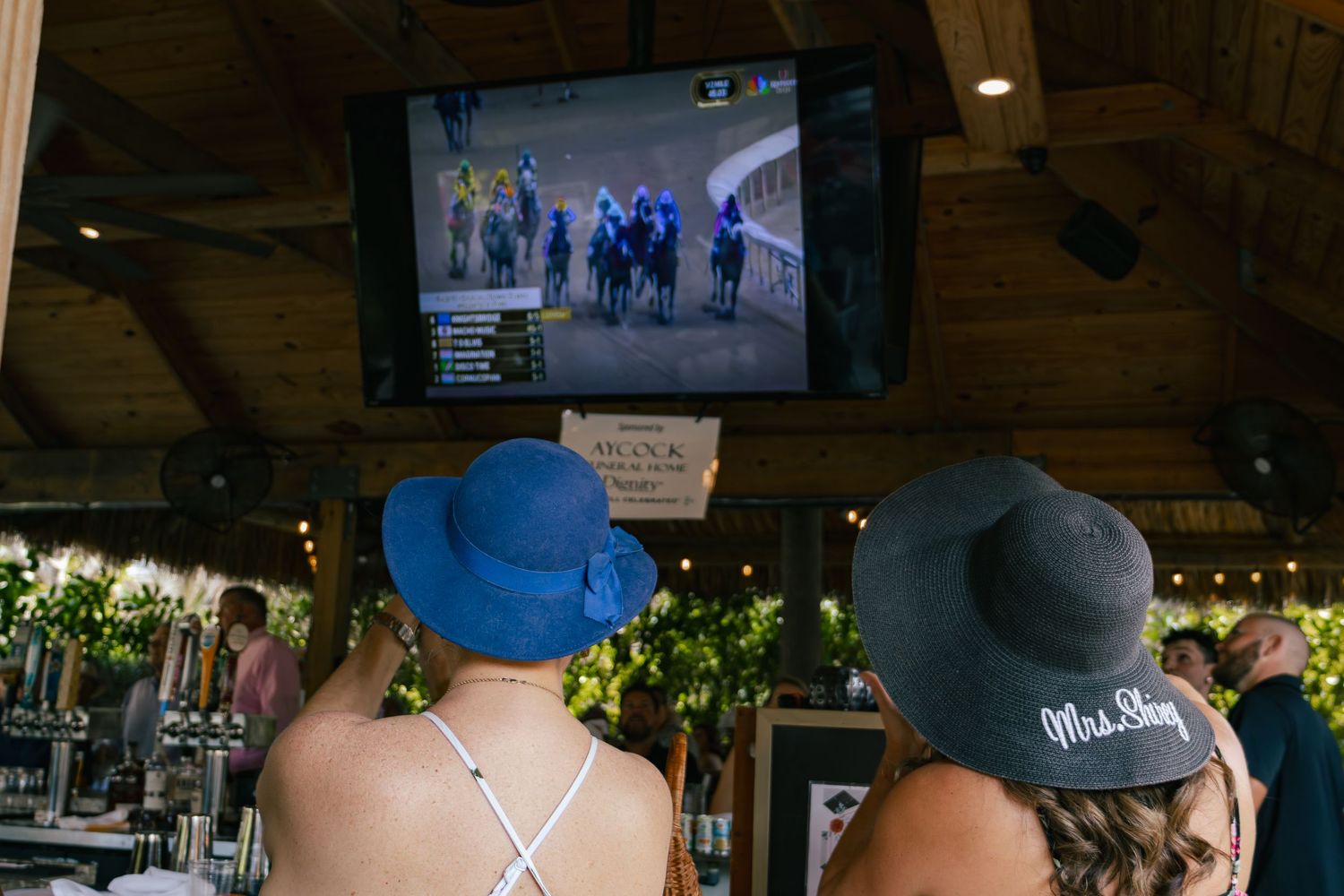 Two women in decorative hats watching horse racing on TV at a bar.