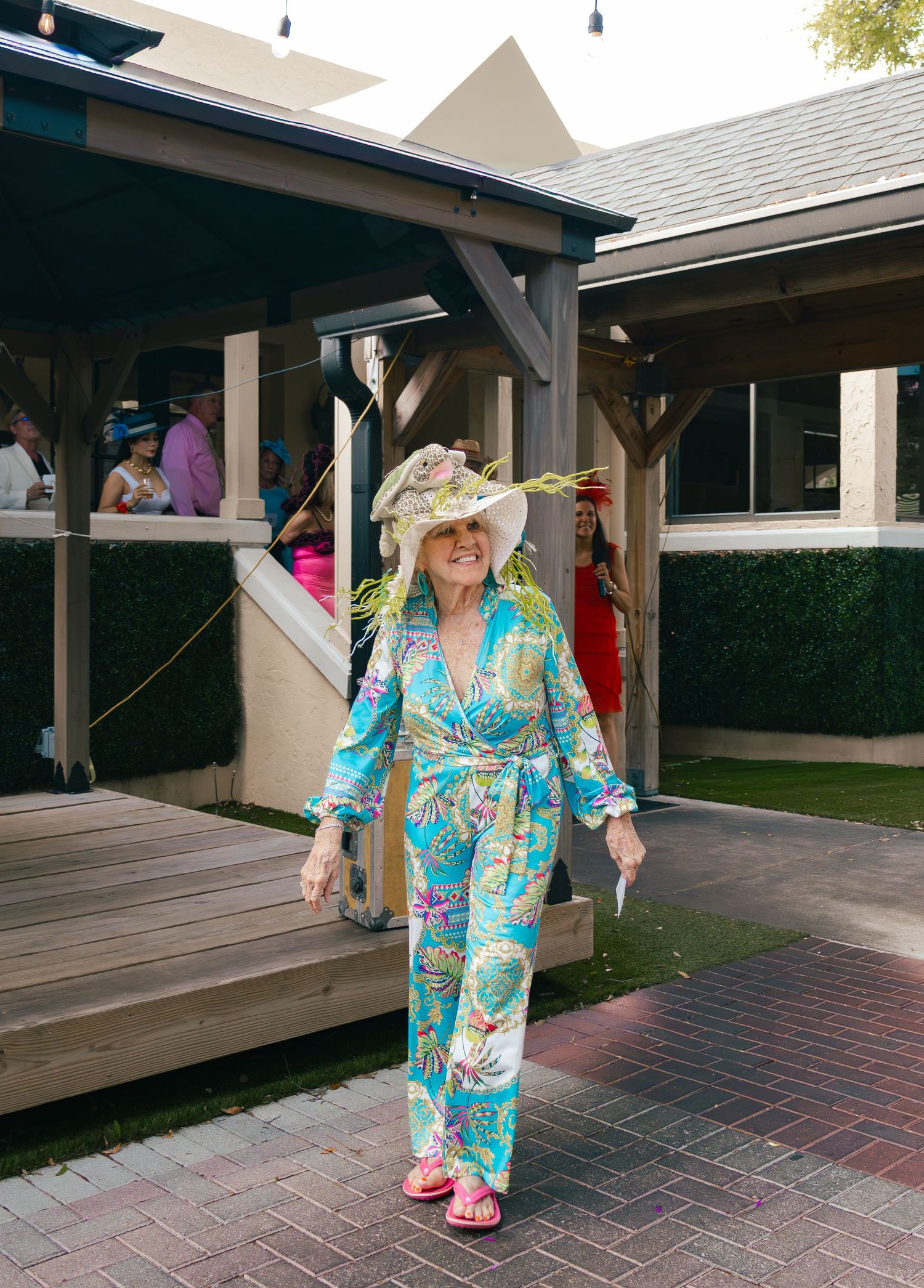 Woman in colorful tropical jumpsuit and floral hat walks at an outdoor Kentucky Derby party event.