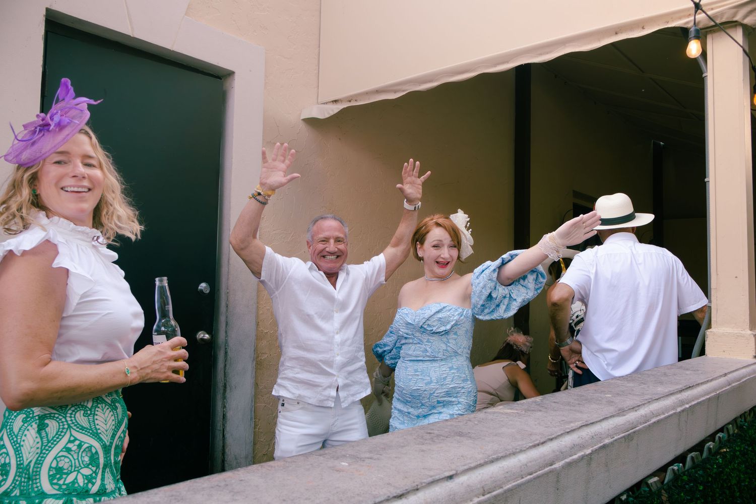 Guests waving from a balcony at an outdoor party, woman in purple hat holding beer bottle.