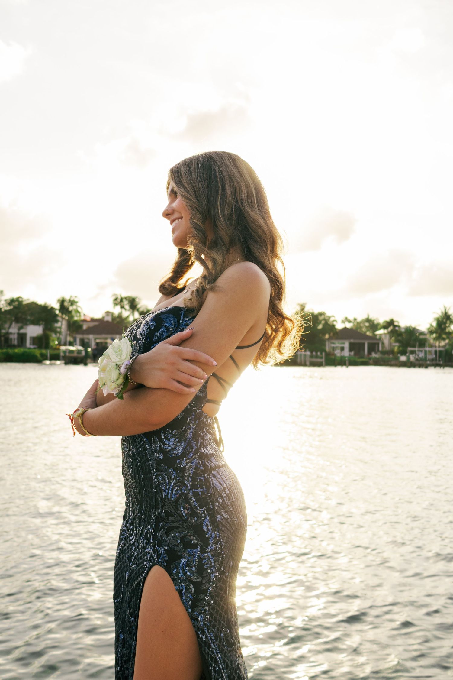 Smiling young woman in dark sequin gown holds white roses while posing by sunlit waterfront.
