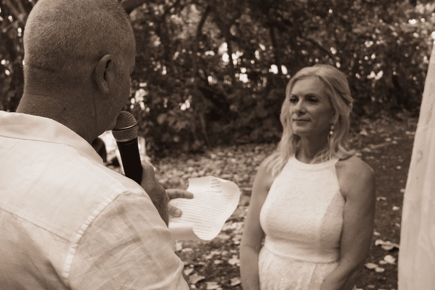 Sepia-toned photo of a groom reading vows to his smiling bride during an outdoor wedding ceremony.