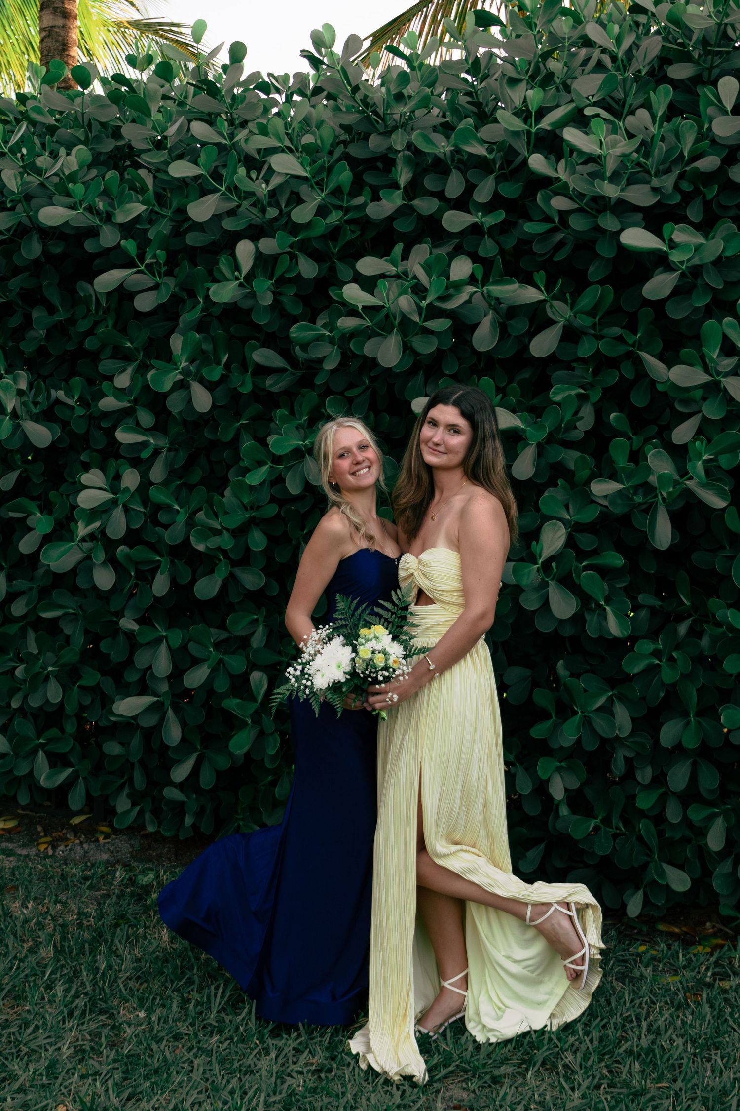 Two smiling women in navy and yellow formal gowns pose together before green hedge backdrop.