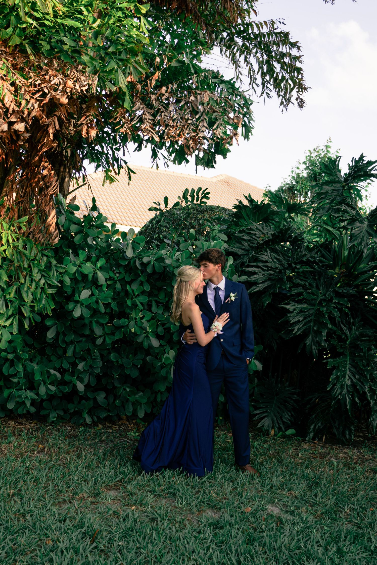 Couple sharing a romantic kiss in a lush garden, wearing navy blue formal attire for prom.