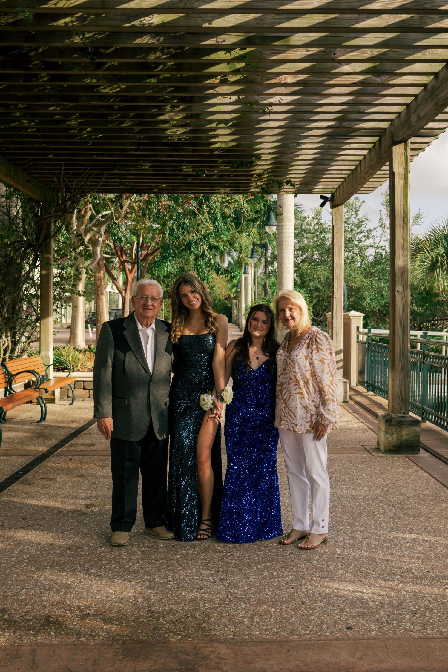 Family of four posing under a pergola with two young women in elegant sequin prom dresses.