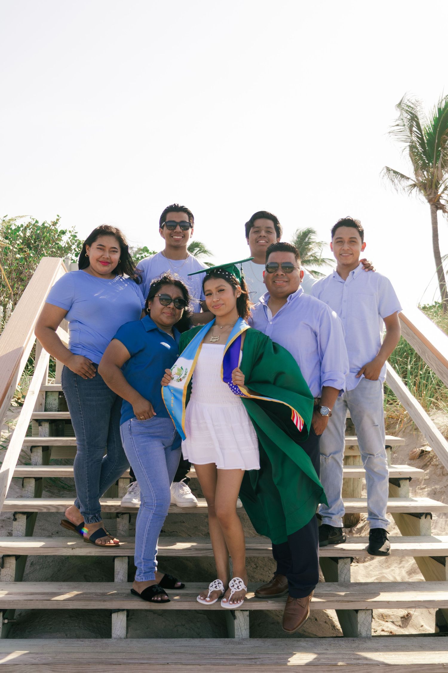 Family of seven poses together on wooden beach stairs celebrating a graduation in green cap and gown.