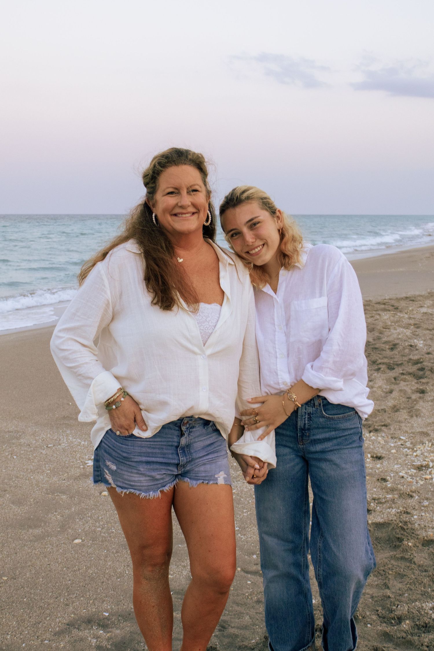 Two women in white shirts smiling together on a beach at dusk with ocean waves behind them.