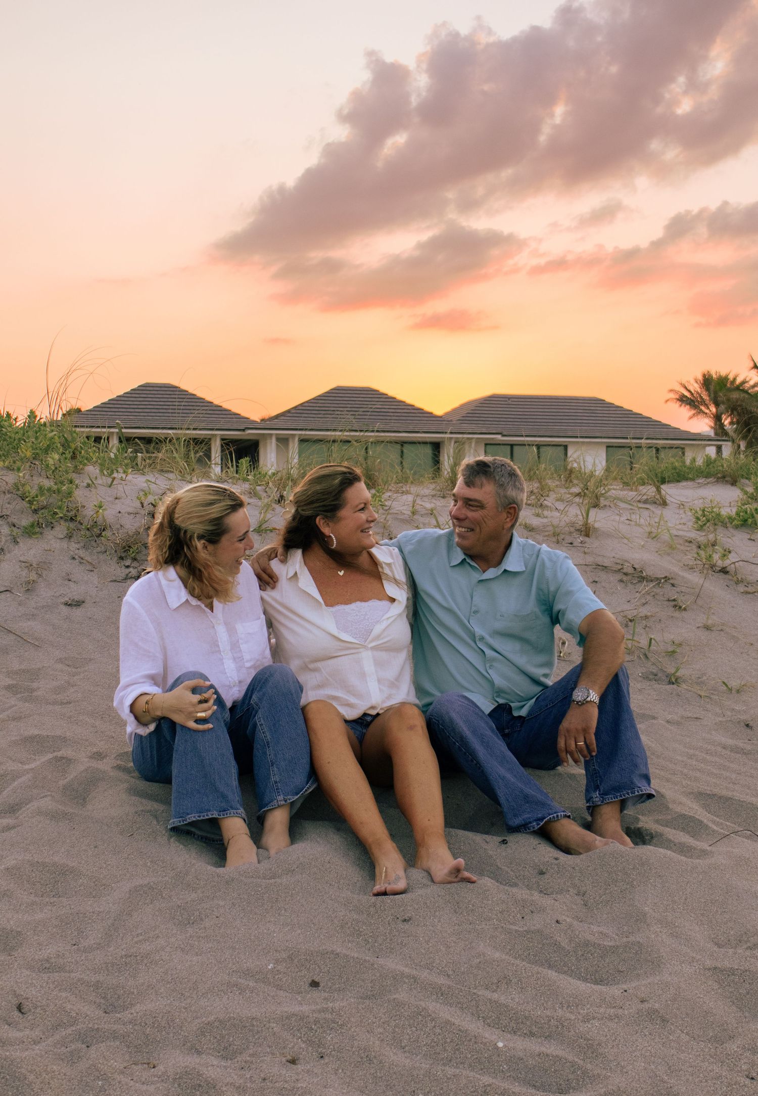 Three people sitting on sandy beach at sunset, smiling and laughing together near beachfront homes.