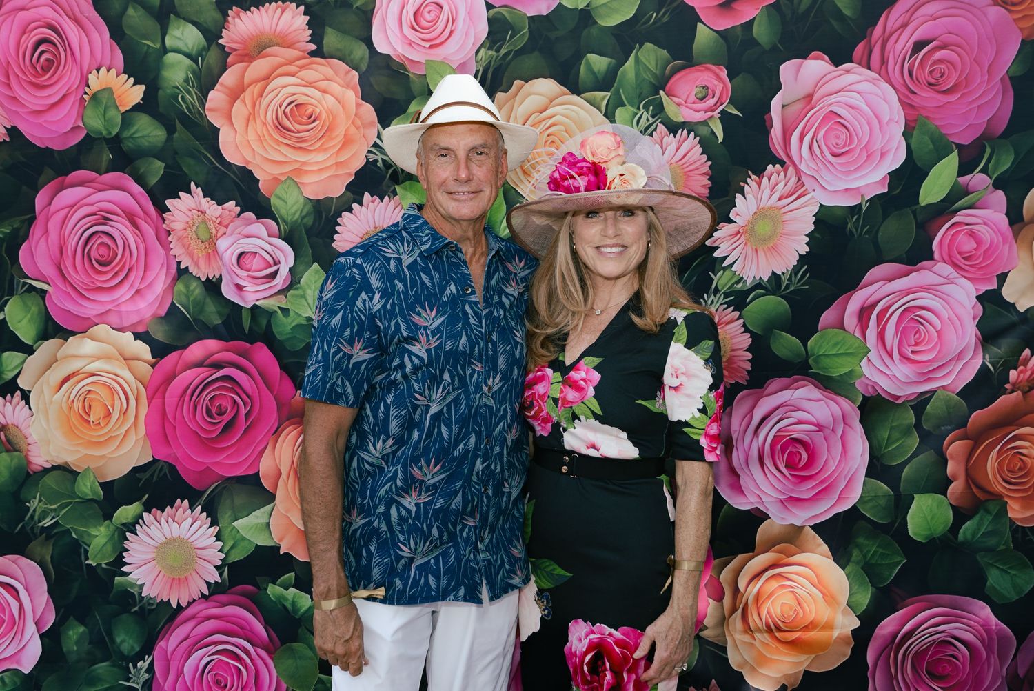Couple posing together in front of a colorful floral rose backdrop at a Kentucky Derby themed party.