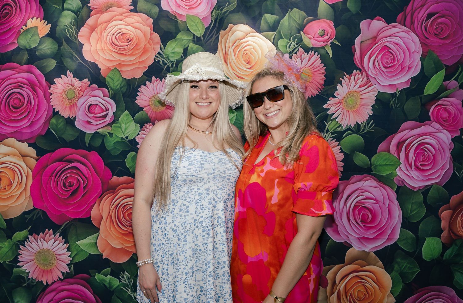 Two blonde women in summer dresses and hats posing before a vibrant floral rose backdrop.