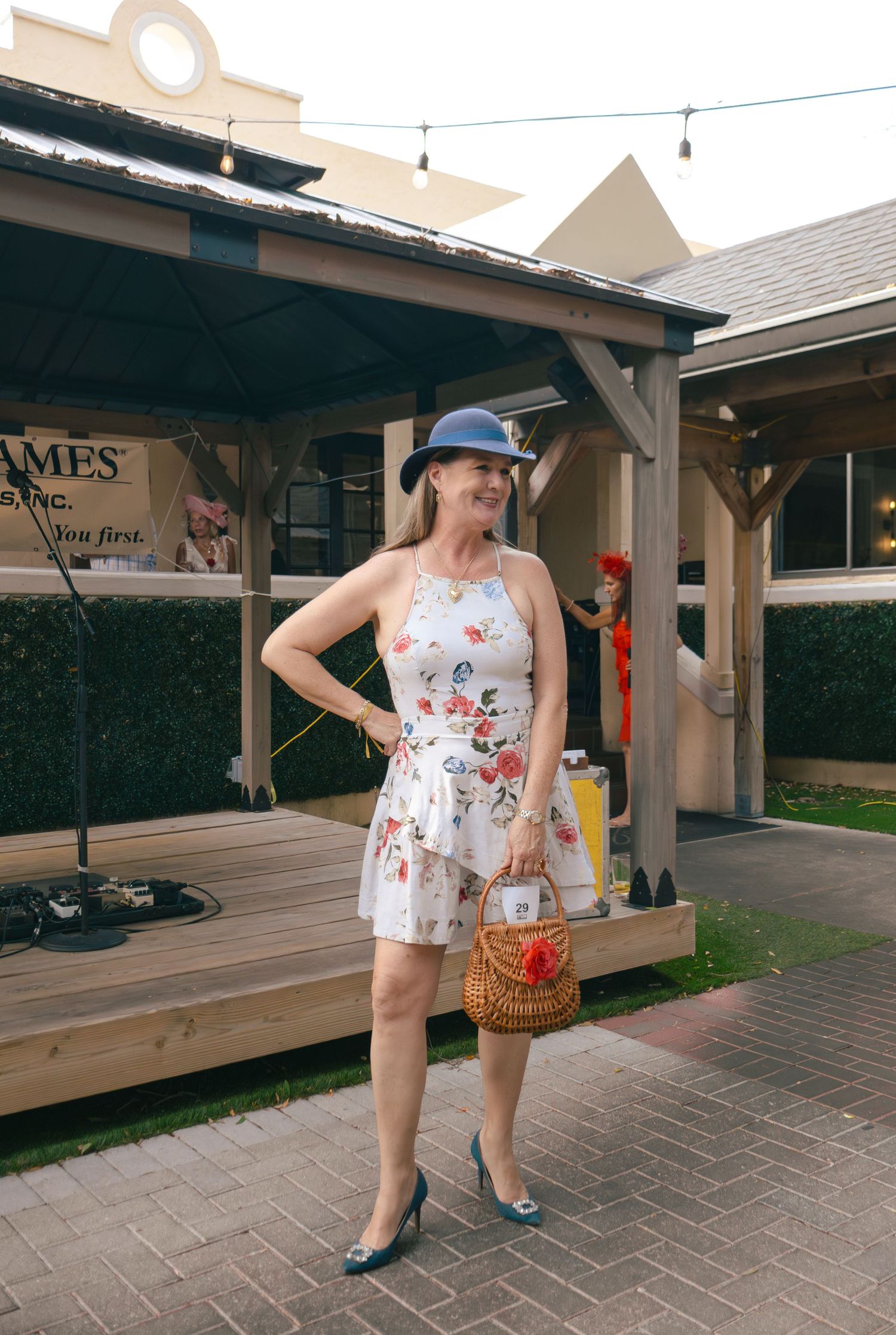 Woman in floral dress and blue hat holds basket purse at outdoor gazebo event.