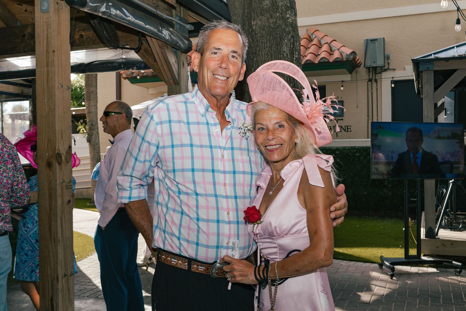 Elegant couple posing outdoors, woman wearing pink fascinator hat and floral dress, man in plaid shirt at social event.