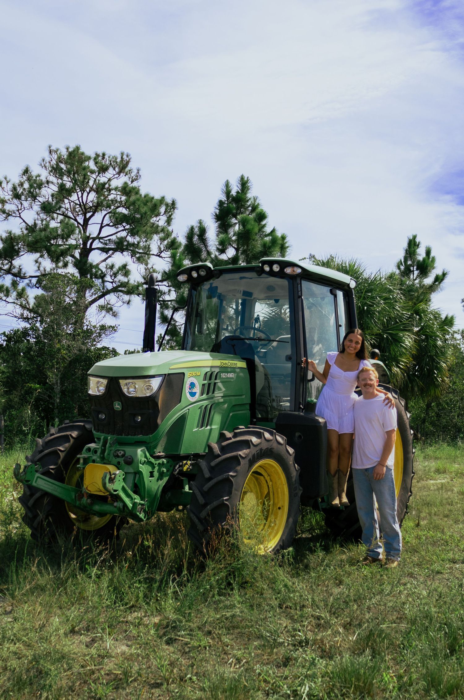 Couple posing in front of a green John Deere tractor in a grassy field with pine trees.