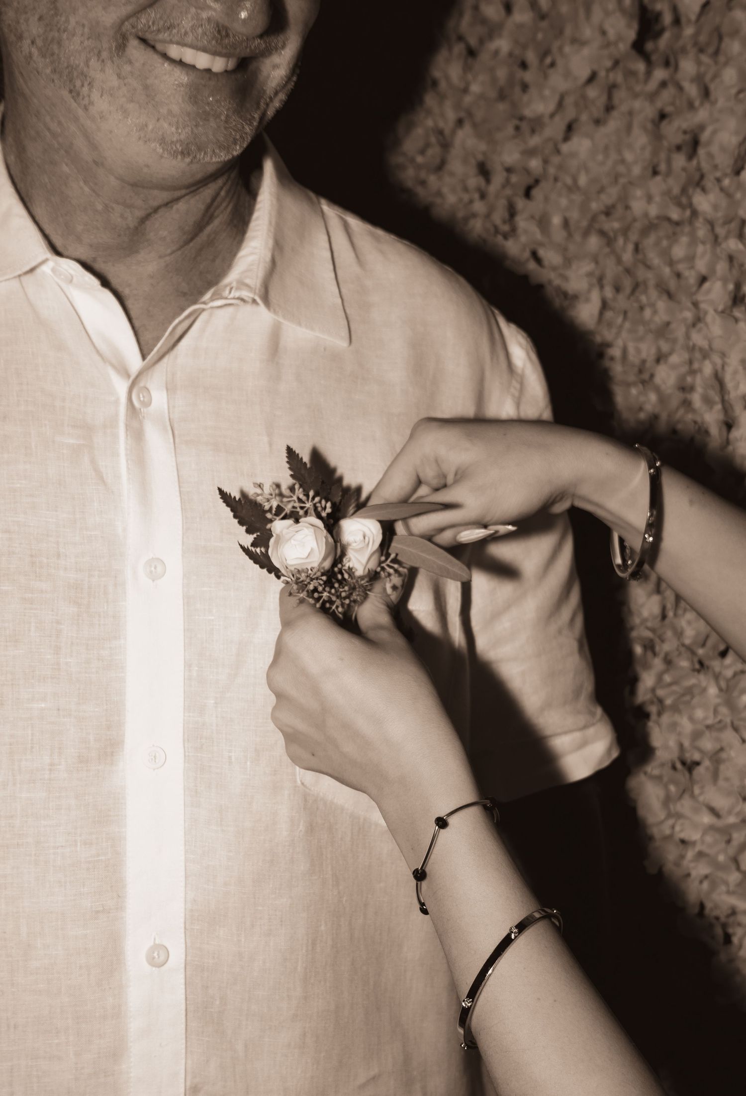 Close-up sepia photo of hands pinning floral boutonniere on groom's white linen shirt.