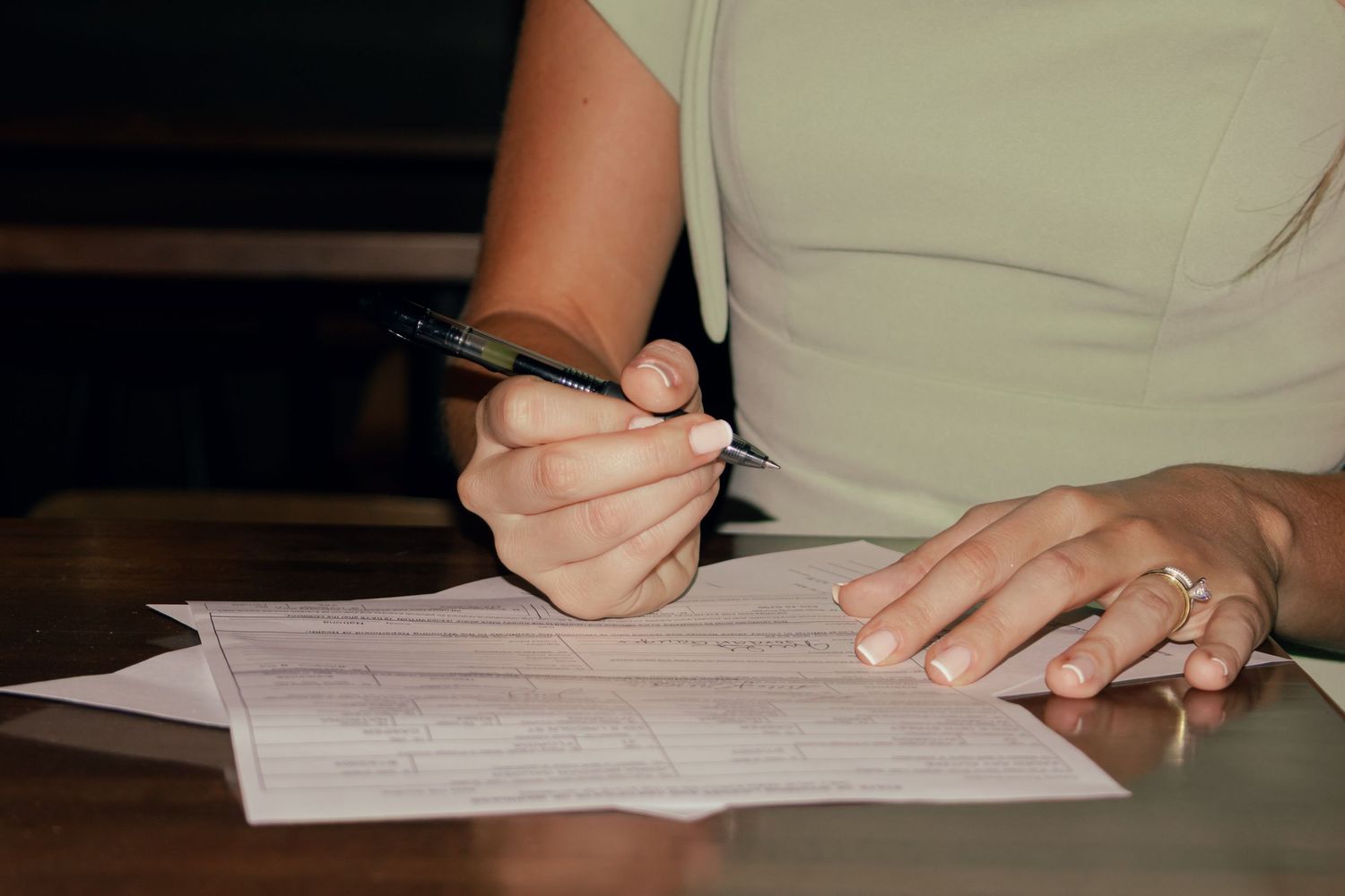 Woman in sage green dress signing wedding documents at wooden table.