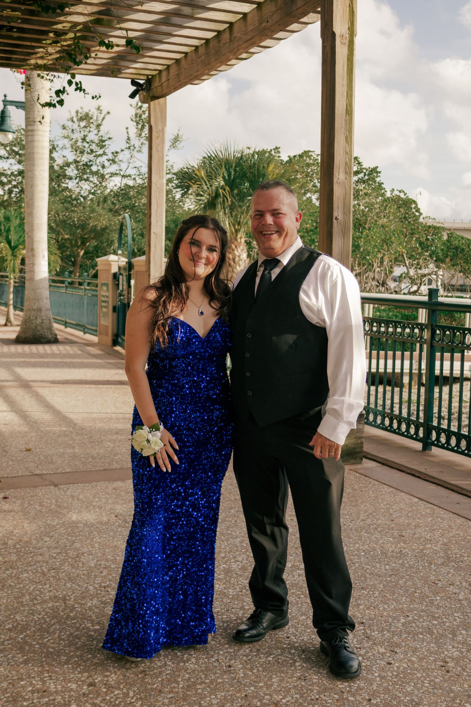 Young woman in a royal blue sequin prom dress posing with a man in a vest under a pergola.