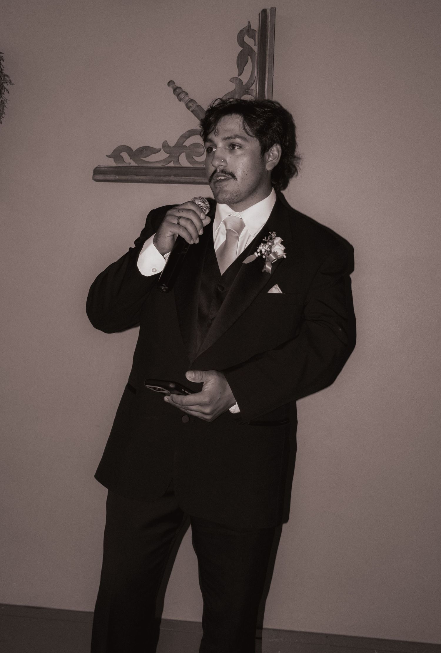 Dapper groomsman in black tuxedo delivering wedding toast with microphone.
