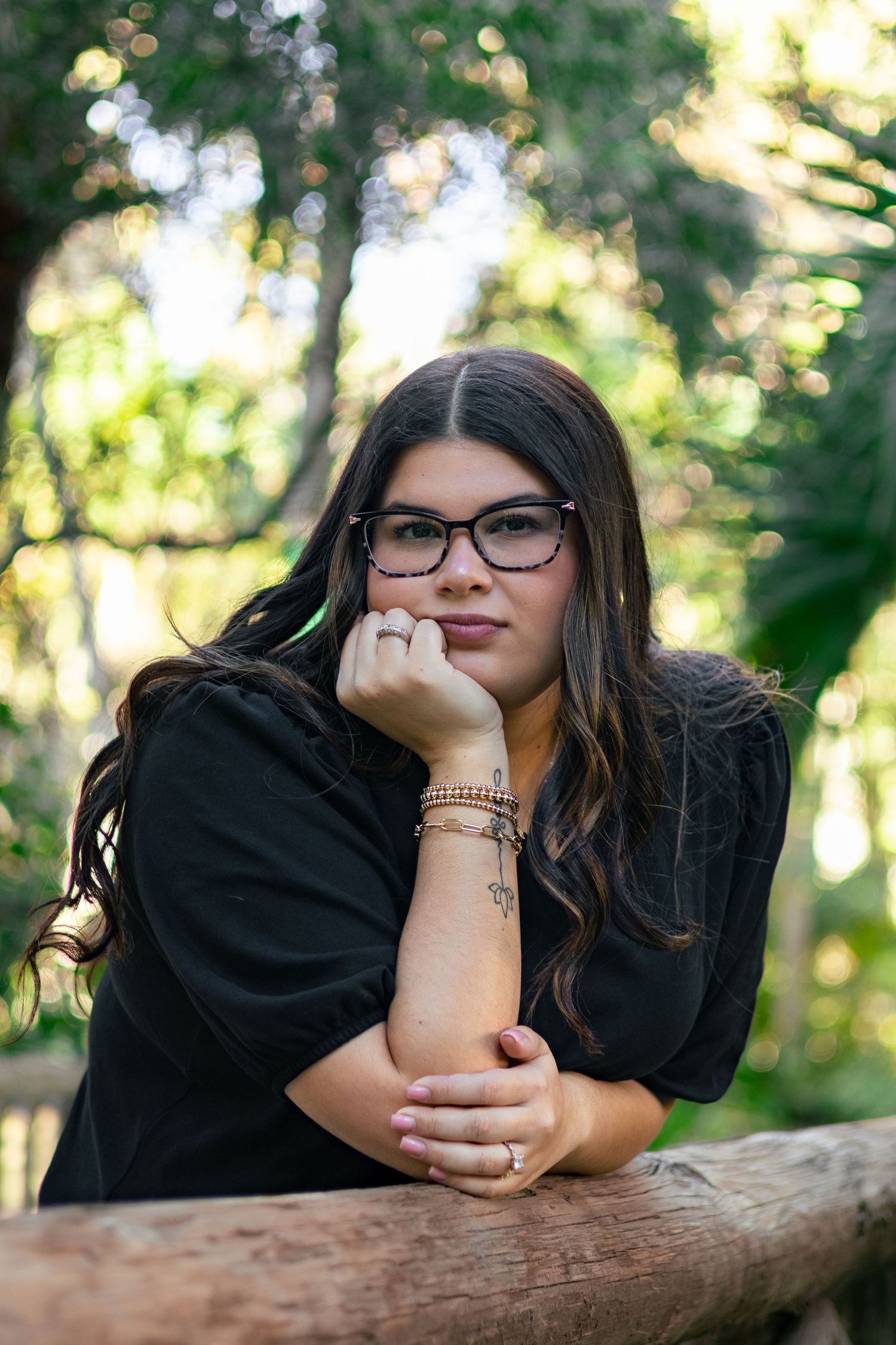 Young woman with glasses and bracelets posing thoughtfully against rustic wooden fence in forest.
