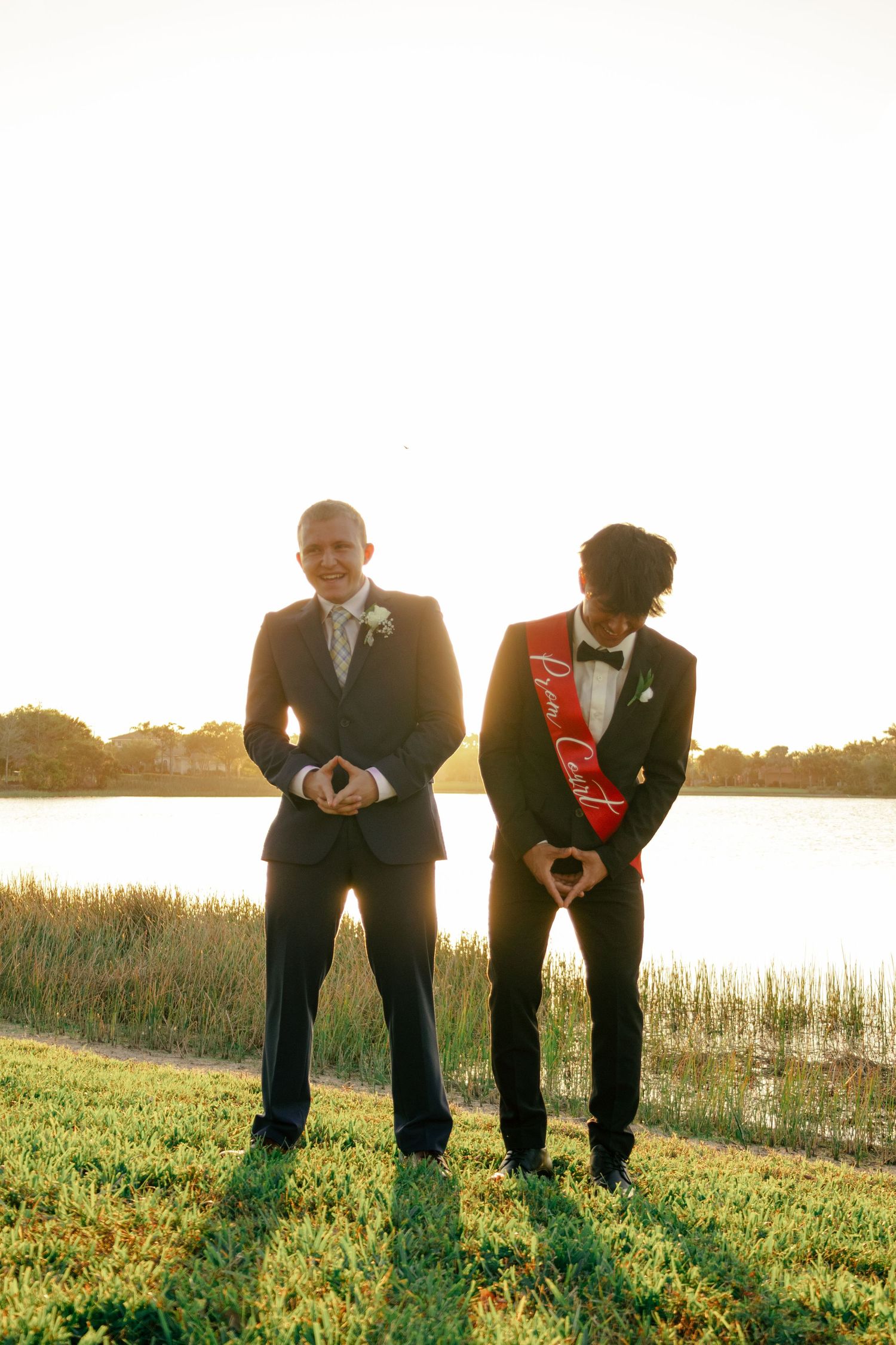 Two young men in dark suits stand outdoors at sunset, one wearing a red Prom Court sash.
