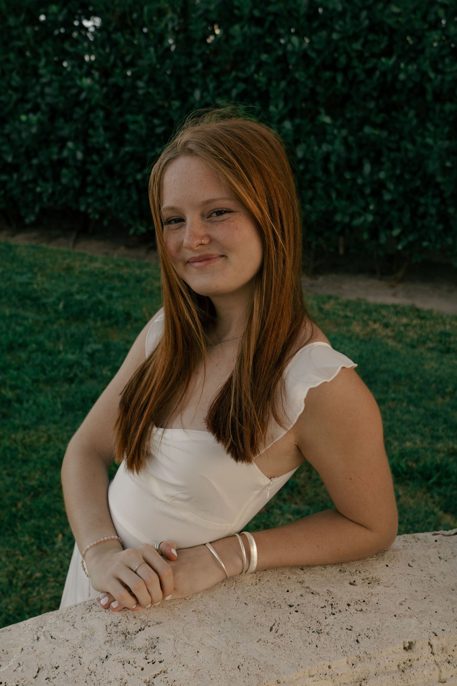 Young woman with long auburn hair smiling in white dress leaning on stone wall with green hedges background.