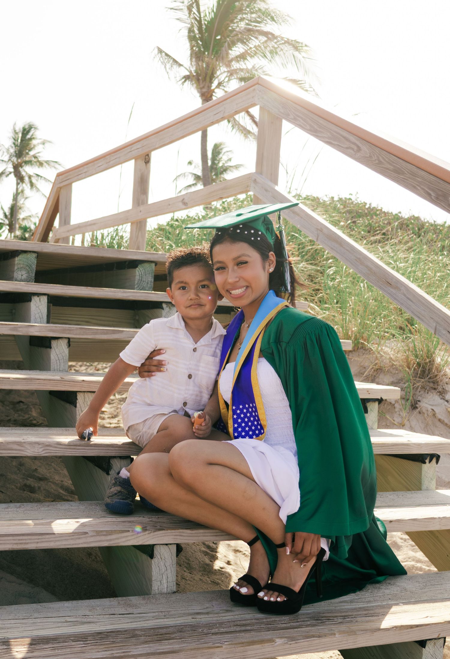 Graduate in green cap and gown sits with young boy on wooden beach stairs with palm trees.