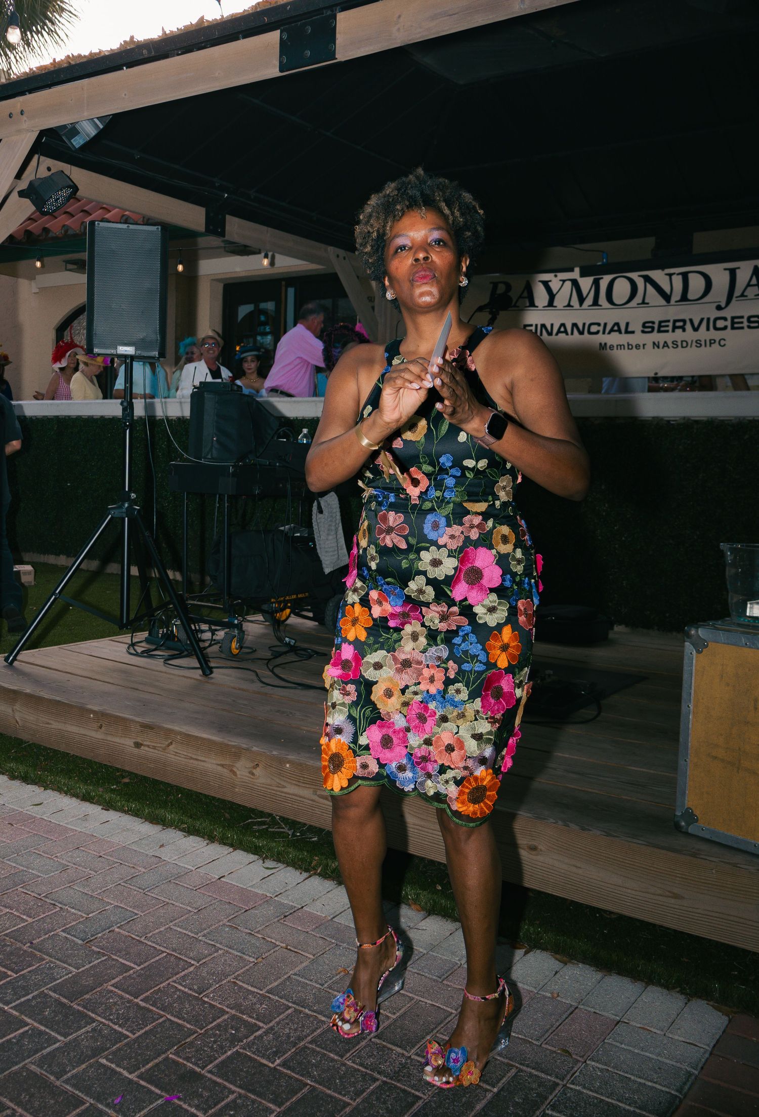 Woman in floral dress clapping at outdoor evening event with live band and Raymond James Financial Services banner.