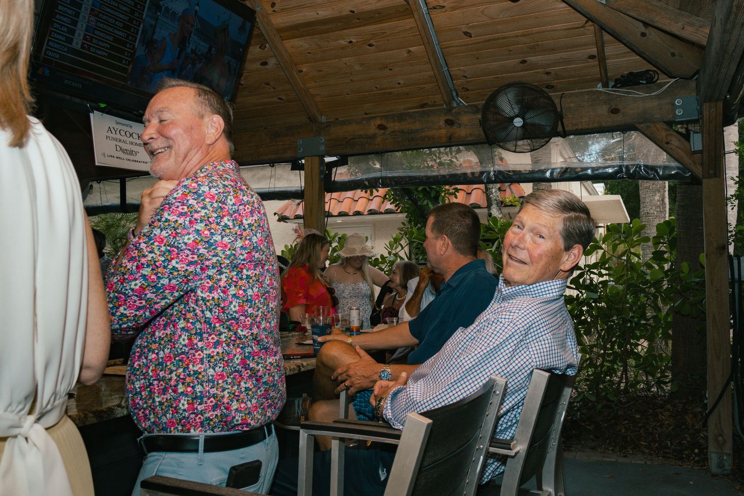 Two men laugh and smile joyfully while seated at an outdoor social gathering event.