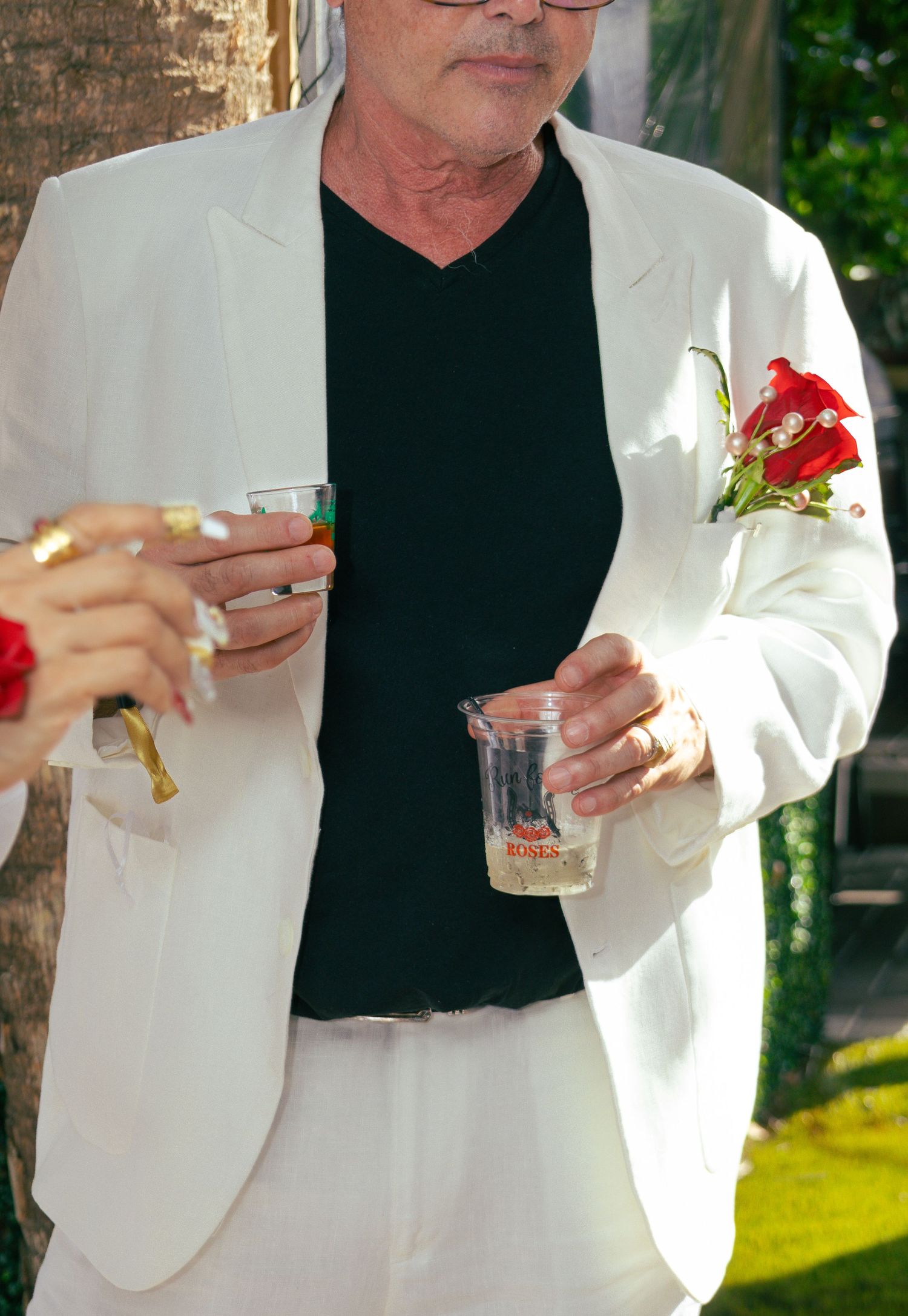 Man in white suit with red rose boutonniere holding drinks at an outdoor celebration.