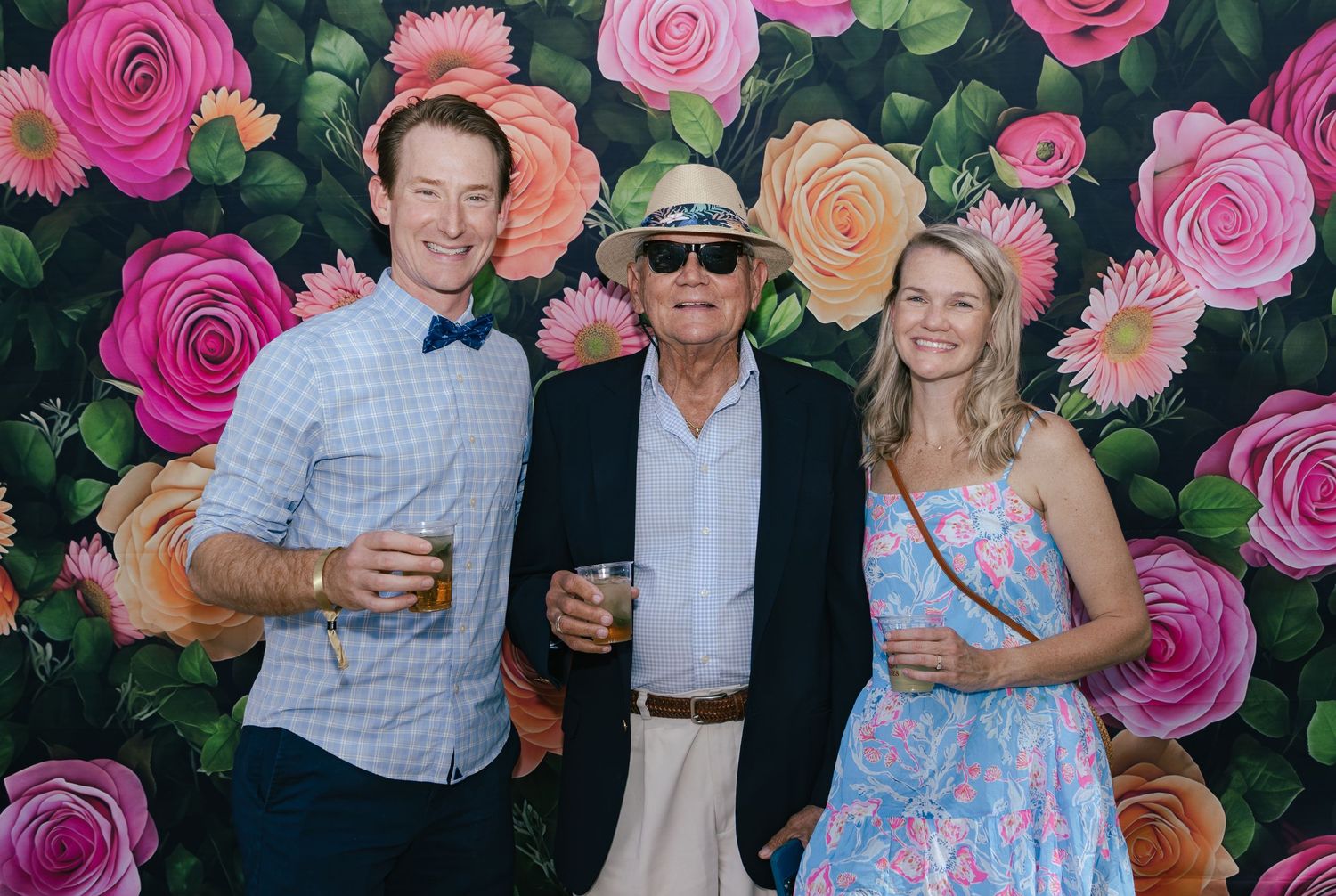 Three people smiling and holding drinks in front of a vibrant floral rose wall backdrop at an event.