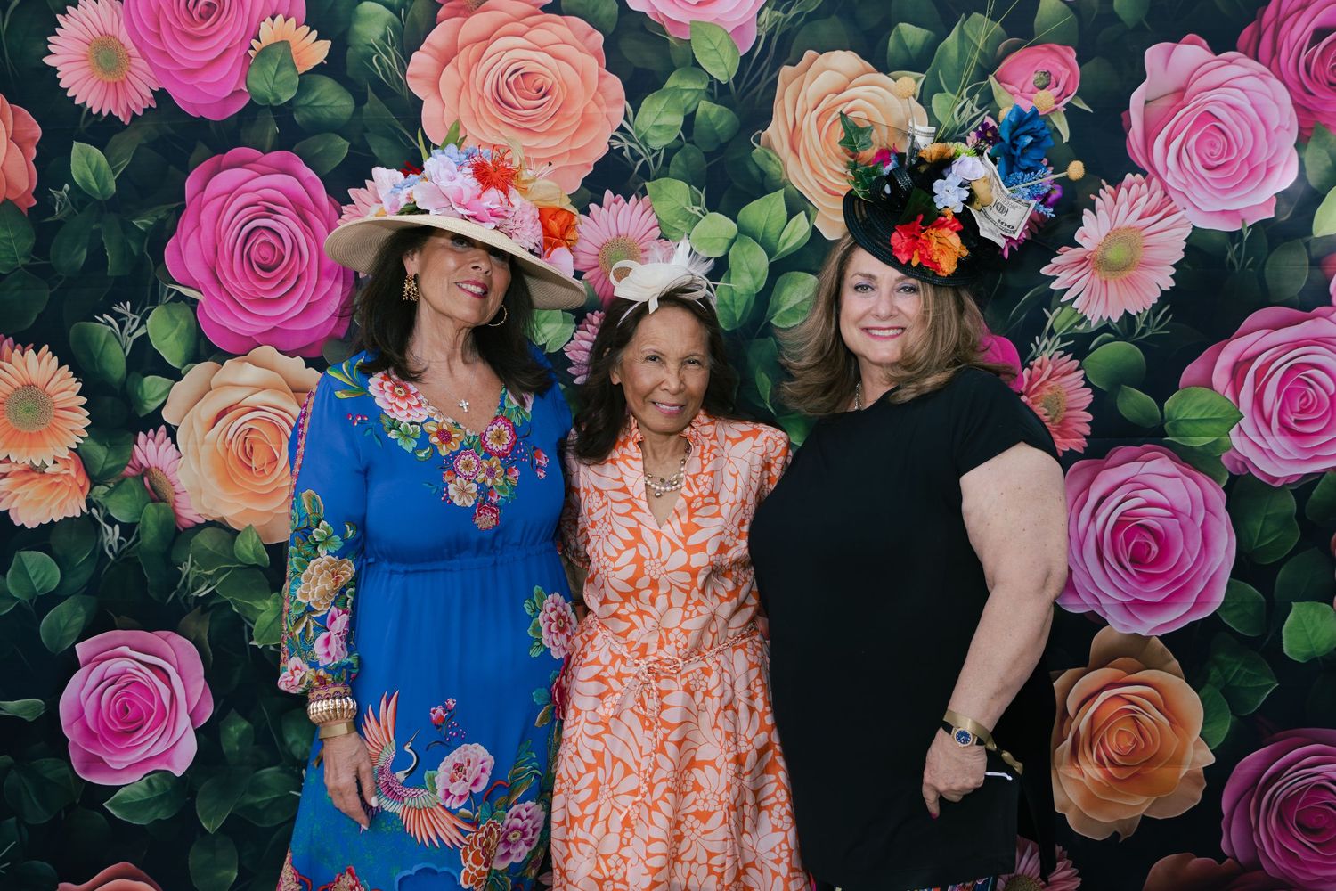 Three women in colorful dresses and decorated hats smiling together in front of a painted floral rose backdrop.
