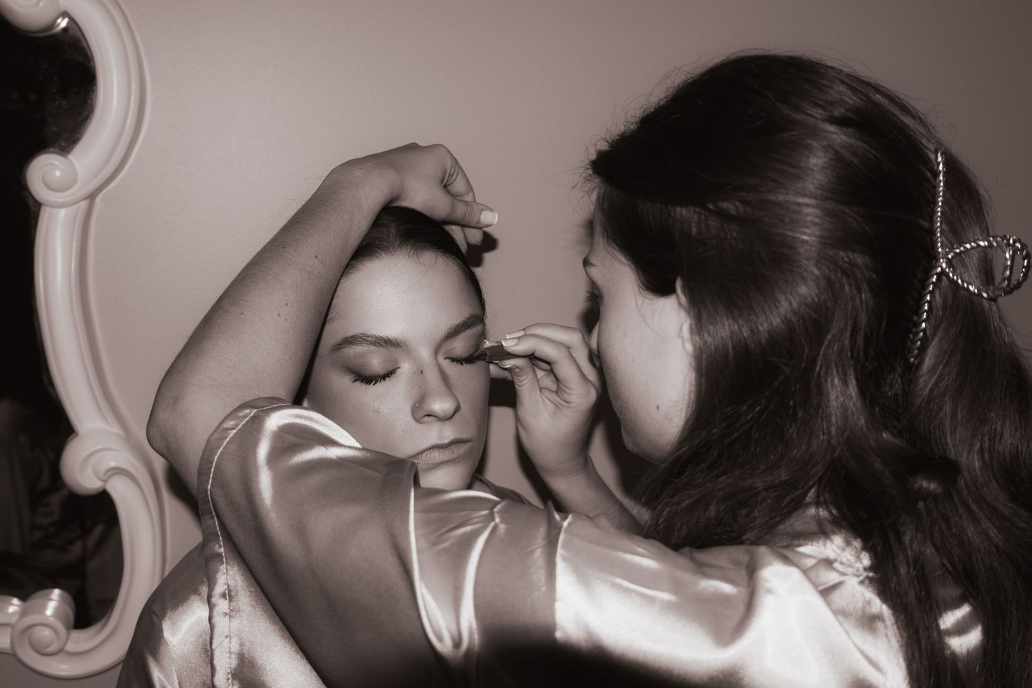 Black and white photo of bridesmaid applying makeup to bride before wedding.