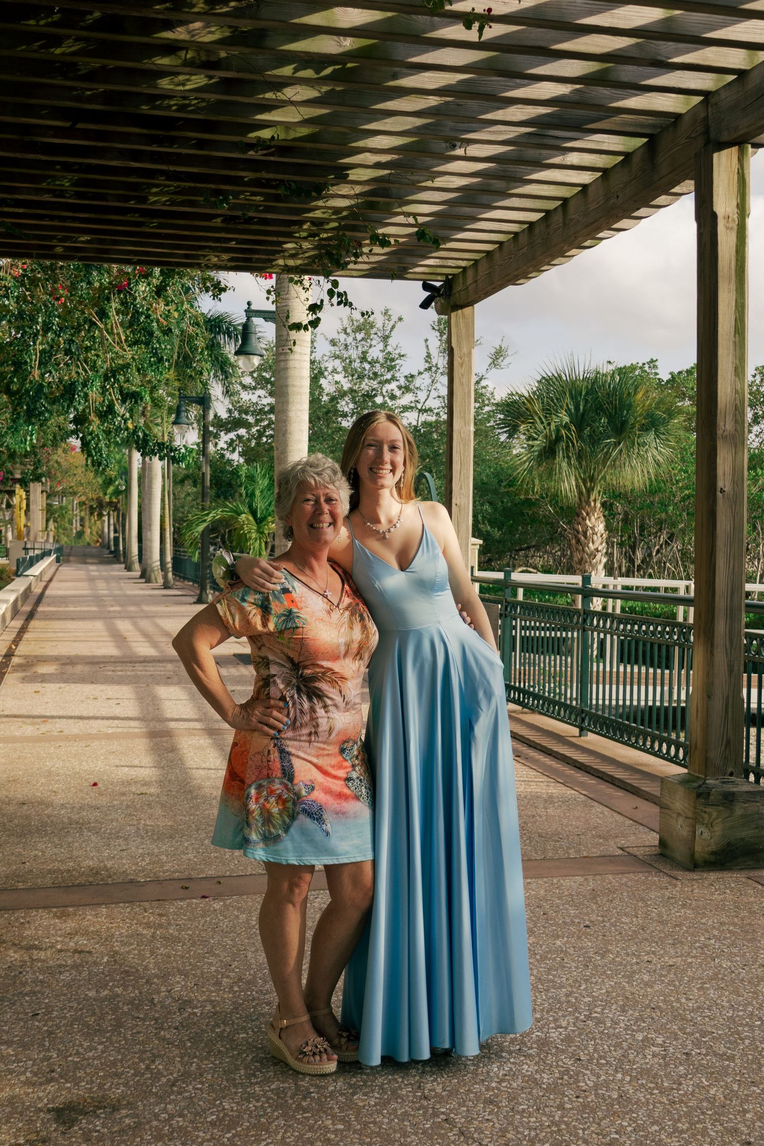 Two women in formal attire pose under a pergola walkway surrounded by lush greenery at a waterfront venue.