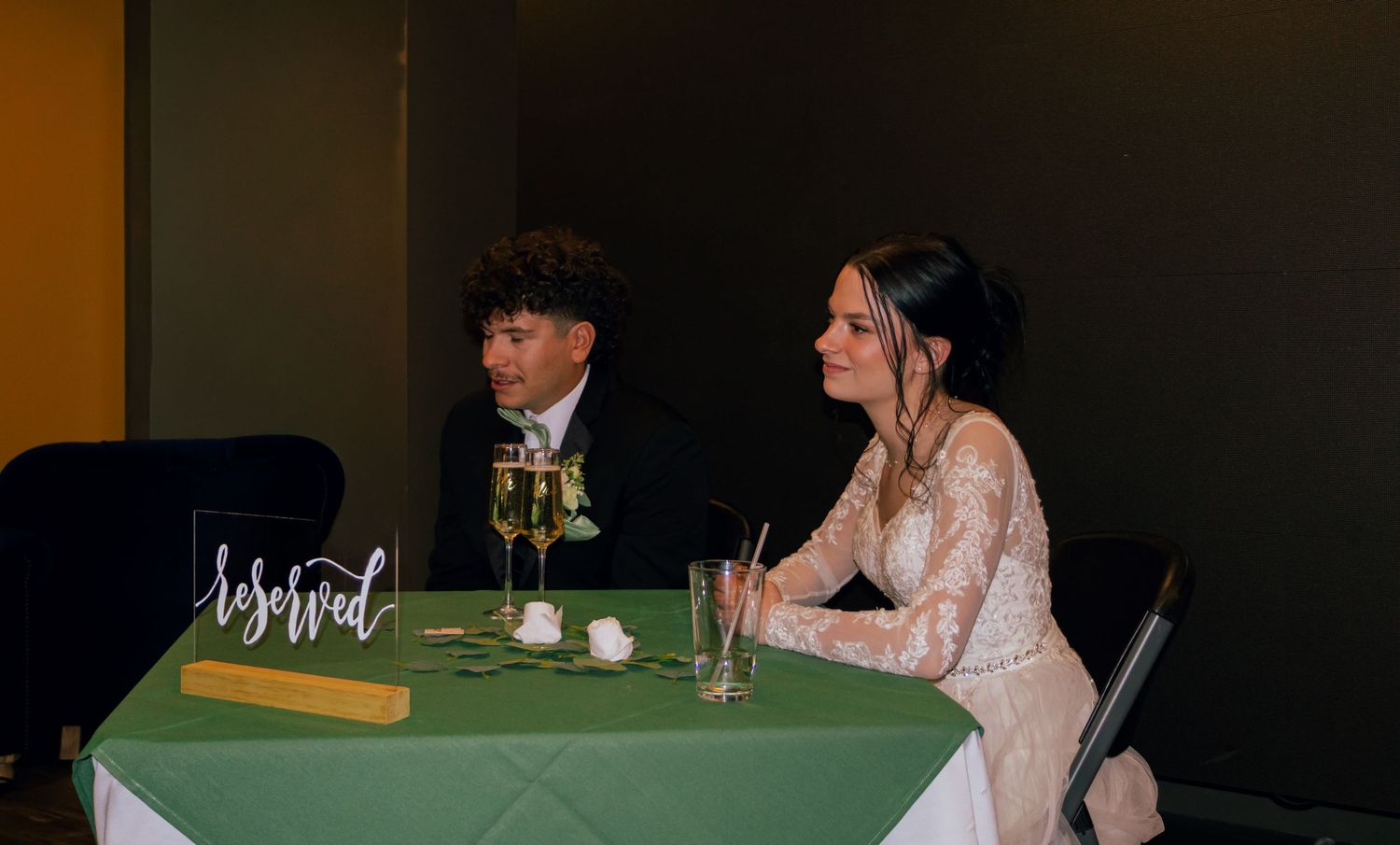 Bride and groom seated at green-draped sweetheart table at wedding reception.
