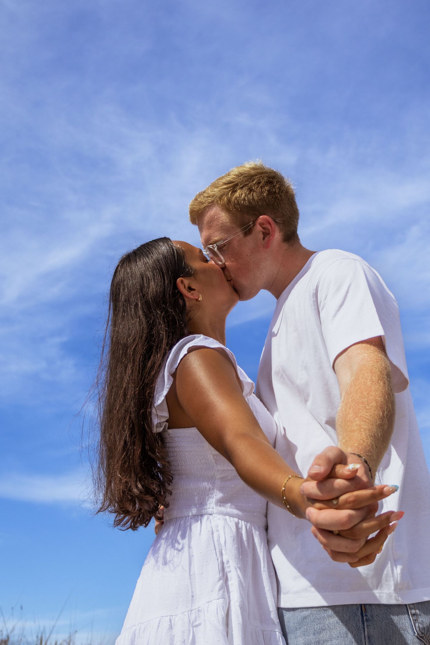 Romantic interracial couple kissing while holding hands against a bright blue sky backdrop.