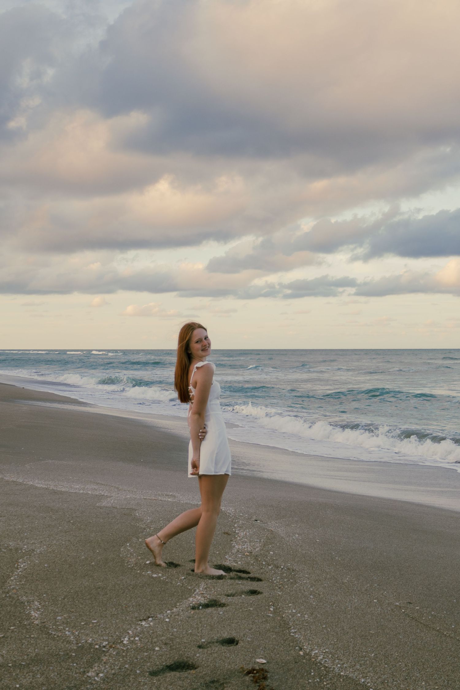 Young woman in white sundress strolling along shoreline at dusk leaving footprints in wet sand.