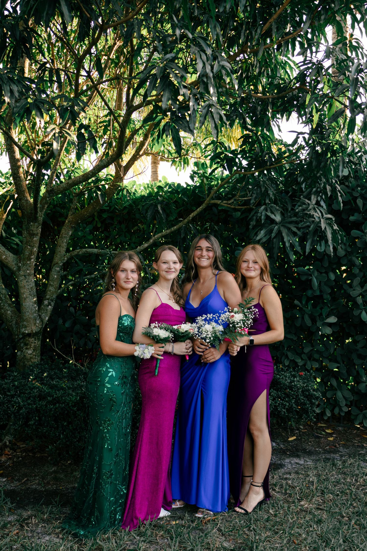 Four smiling young women in colorful prom dresses pose together beneath a lush mango tree.
