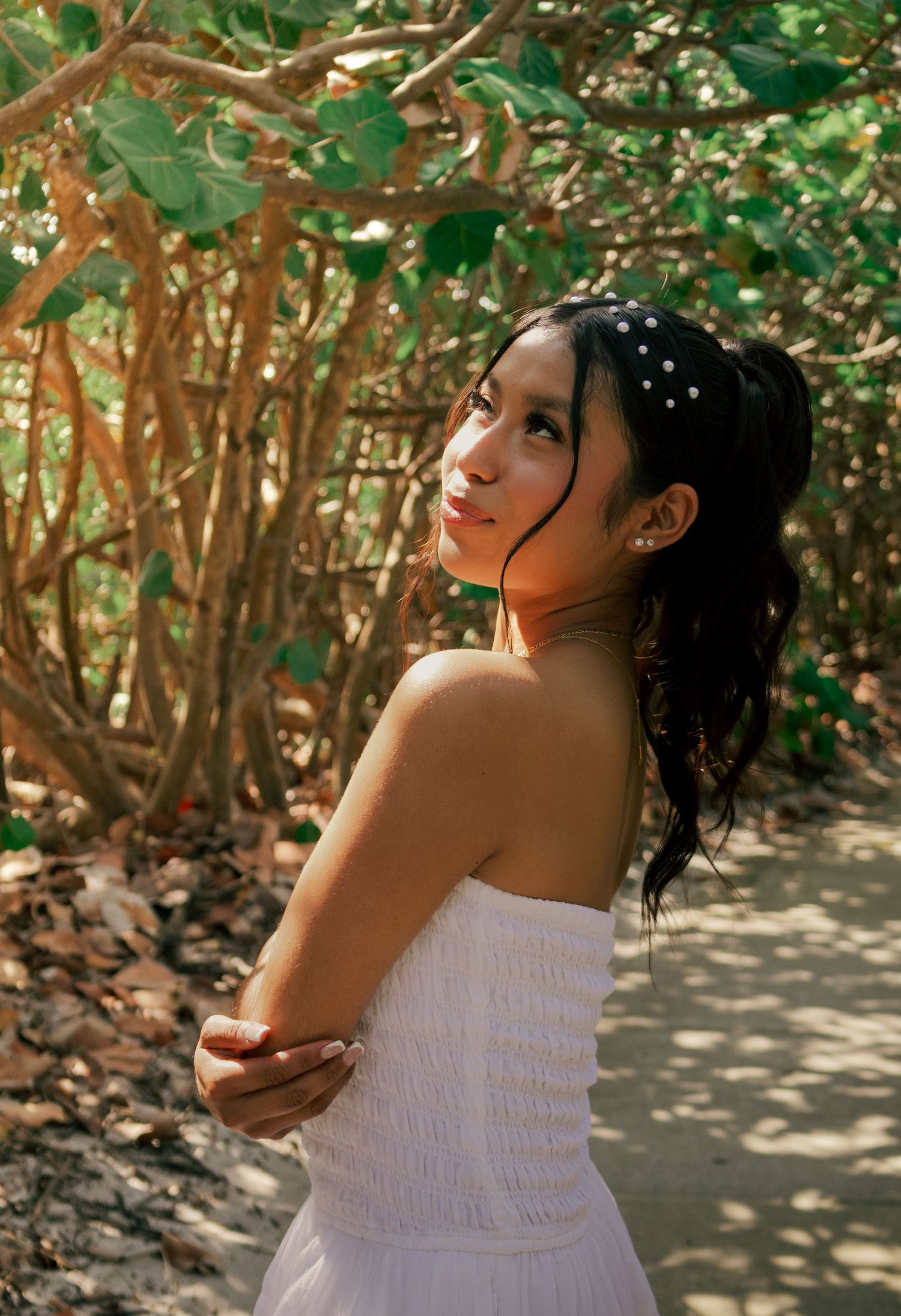 Young woman in white strapless dress with pearl headband looking up gracefully among lush trees in natural light.