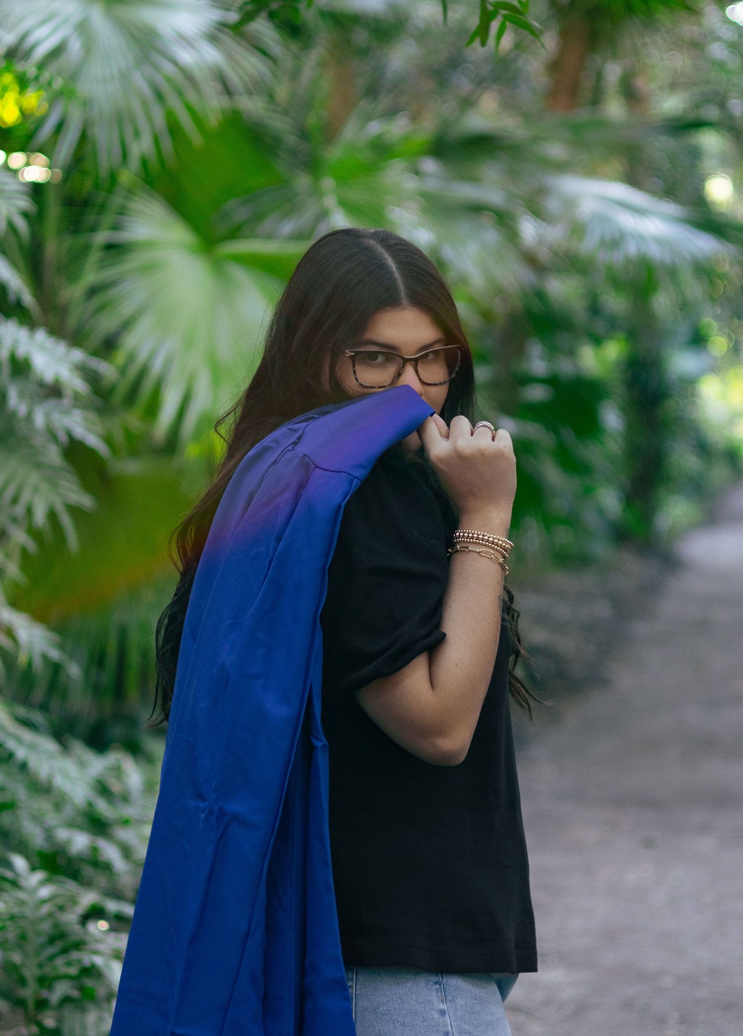 Young woman in black top holding blue graduation gown over shoulder on tropical garden path.
